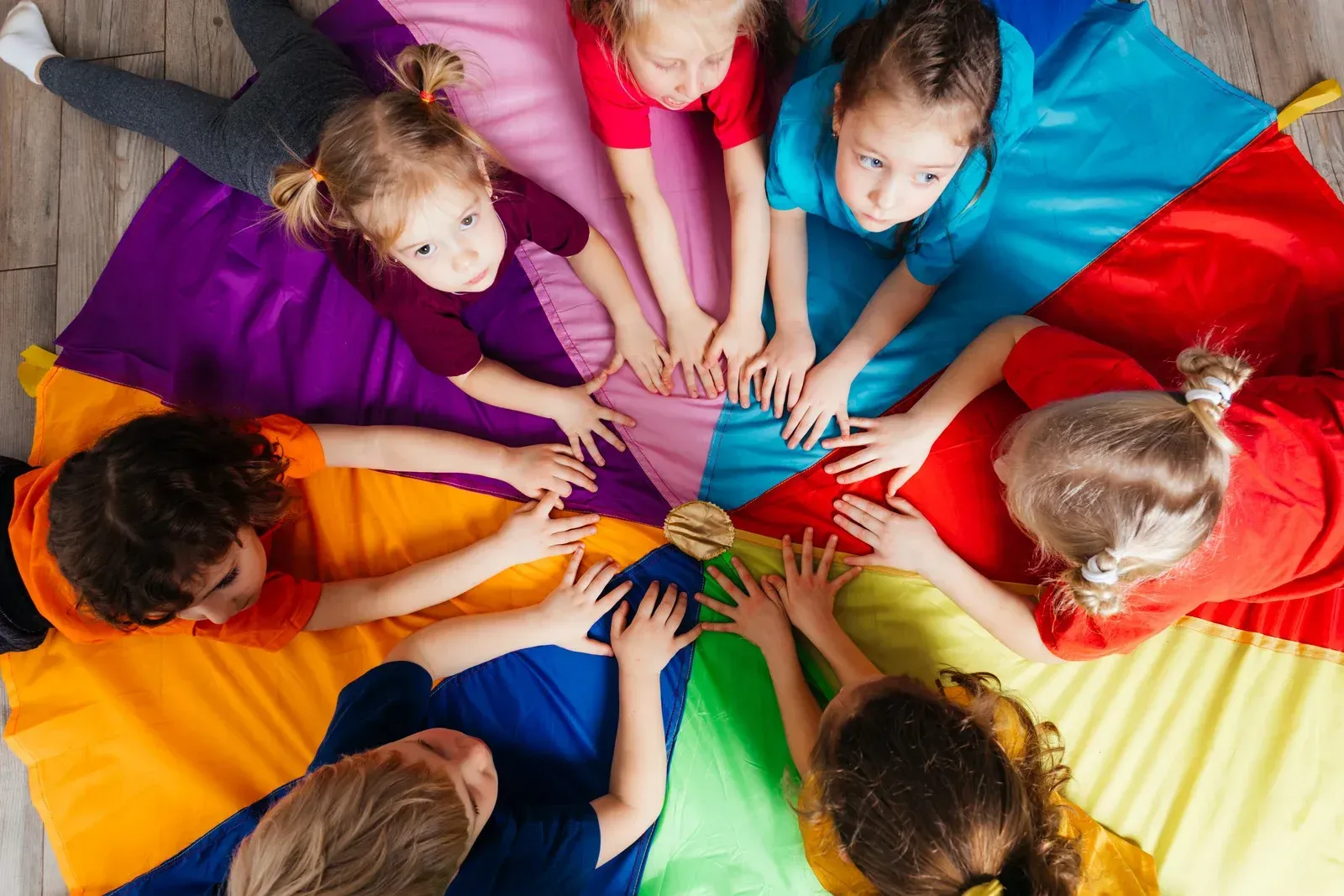 Children gathered around a colorful parachute, hands touching in a circle.