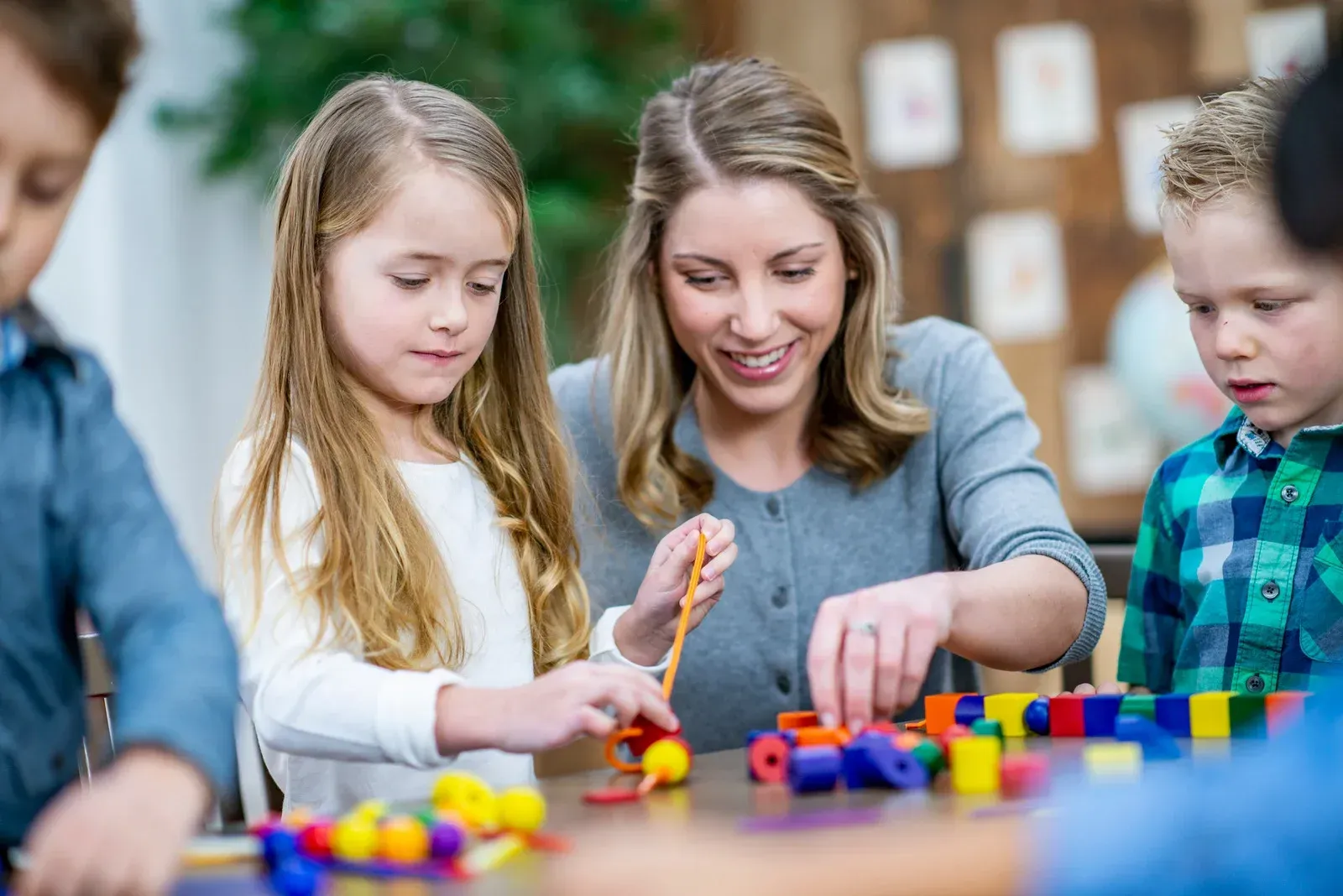A teacher helps children build with colorful blocks at a table in a classroom.