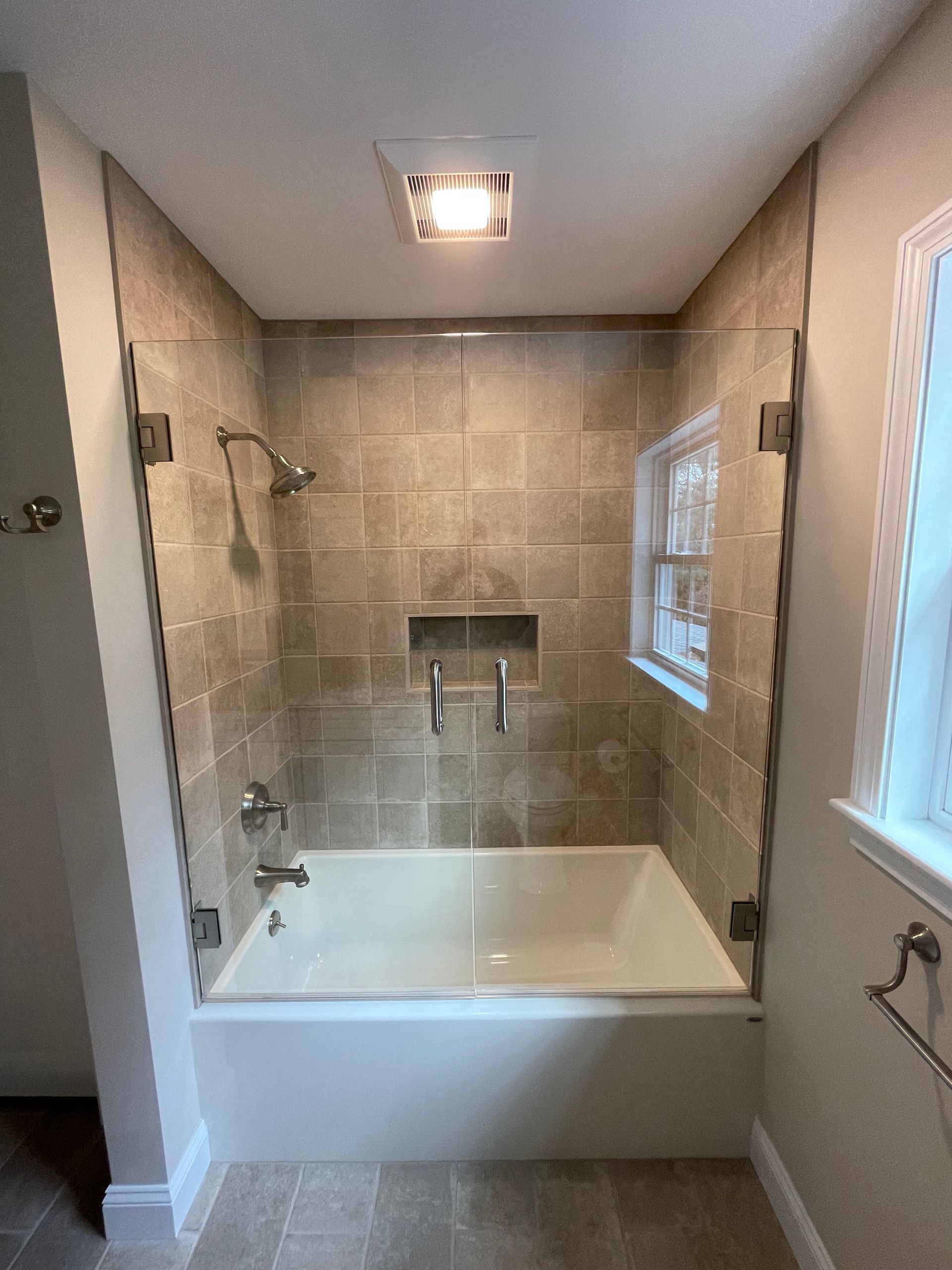 A glass-enclosed bathtub and shower combination featuring beige, square-tiled walls and a built-in recessed shelf.