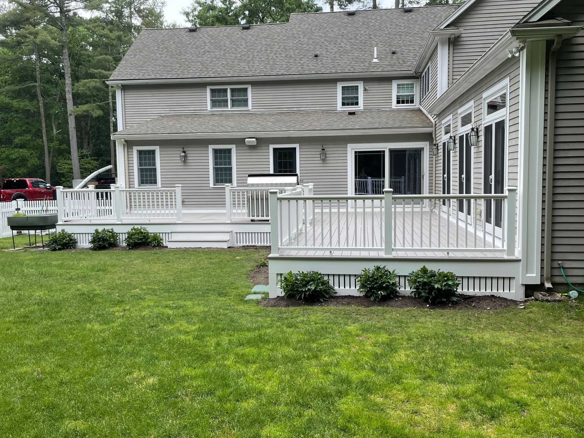 A two-story house with gray siding features a large white deck and railing overlooking a green lawn.