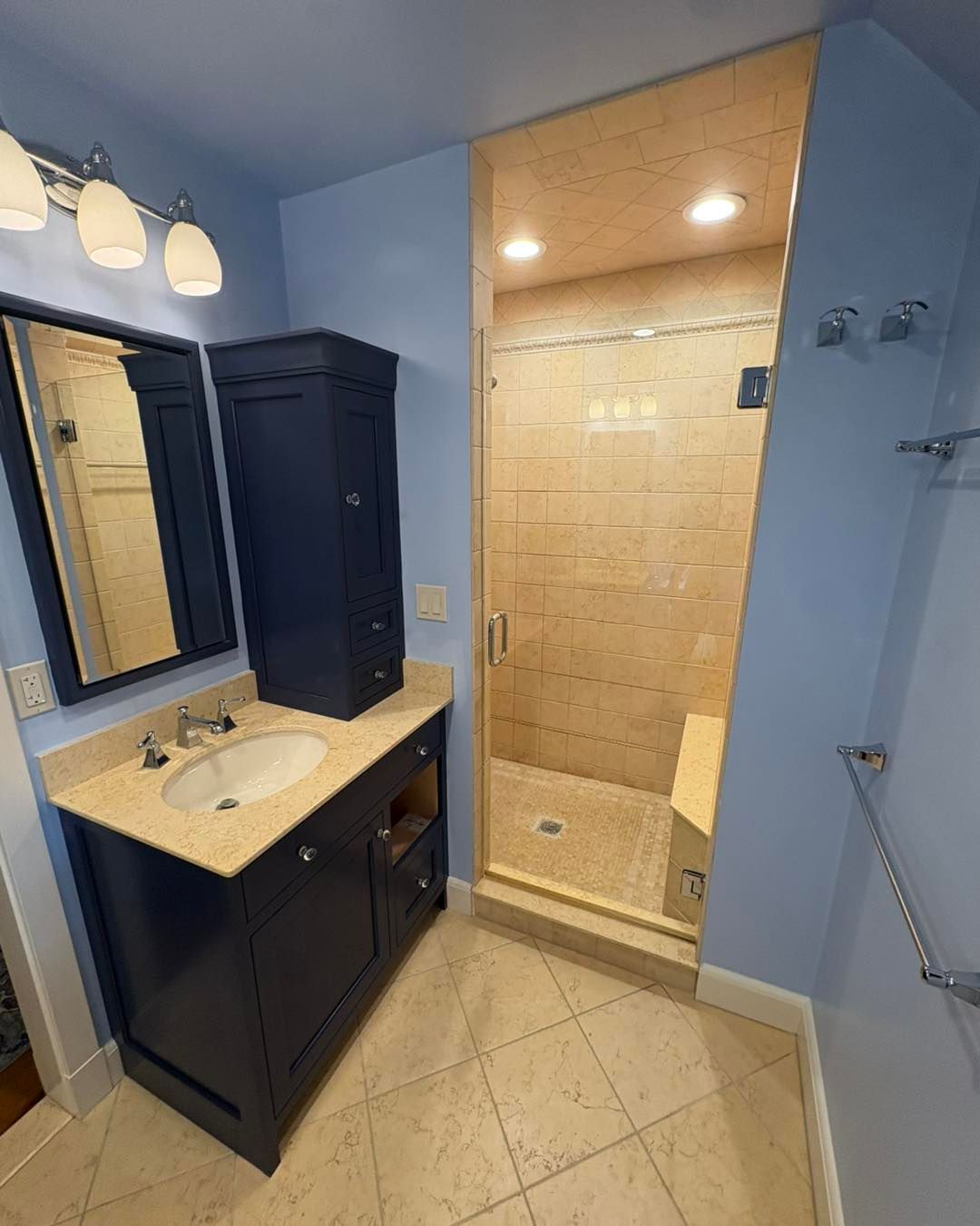 Blue bathroom with navy vanity, tall cabinet, and walk-in shower. Beige tile floors and light blue walls.