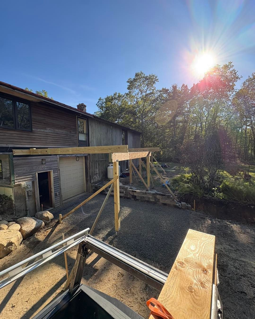Construction of a wooden structure outside a two-story house with gravel ground. Bright sunlight shines through trees.