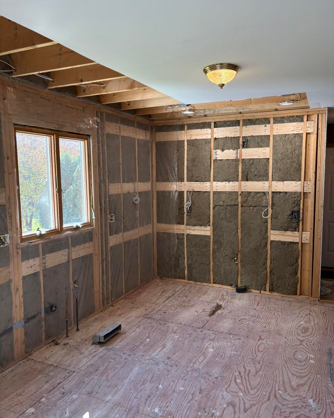 Interior kitchen remodel with exposed studs, insulation, window, light fixture, and plywood floor.
