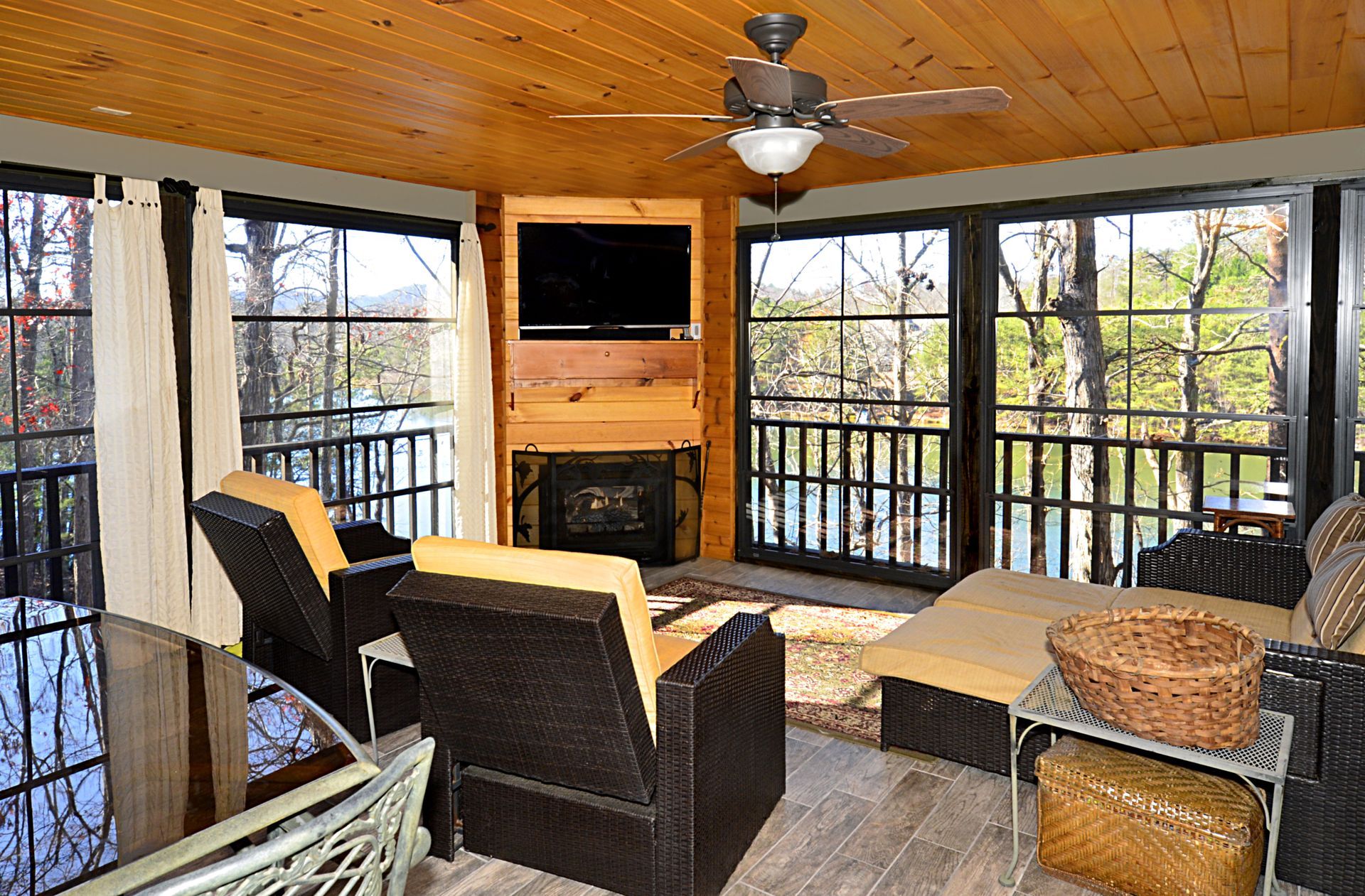 Sunroom with black-framed windows, wicker furniture, fireplace, TV, and view of trees and water.