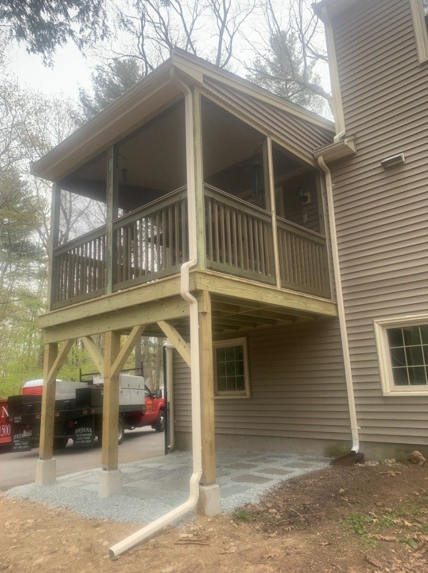 Wooden screened porch with railing and a gabled roof, attached to a light brown house.
