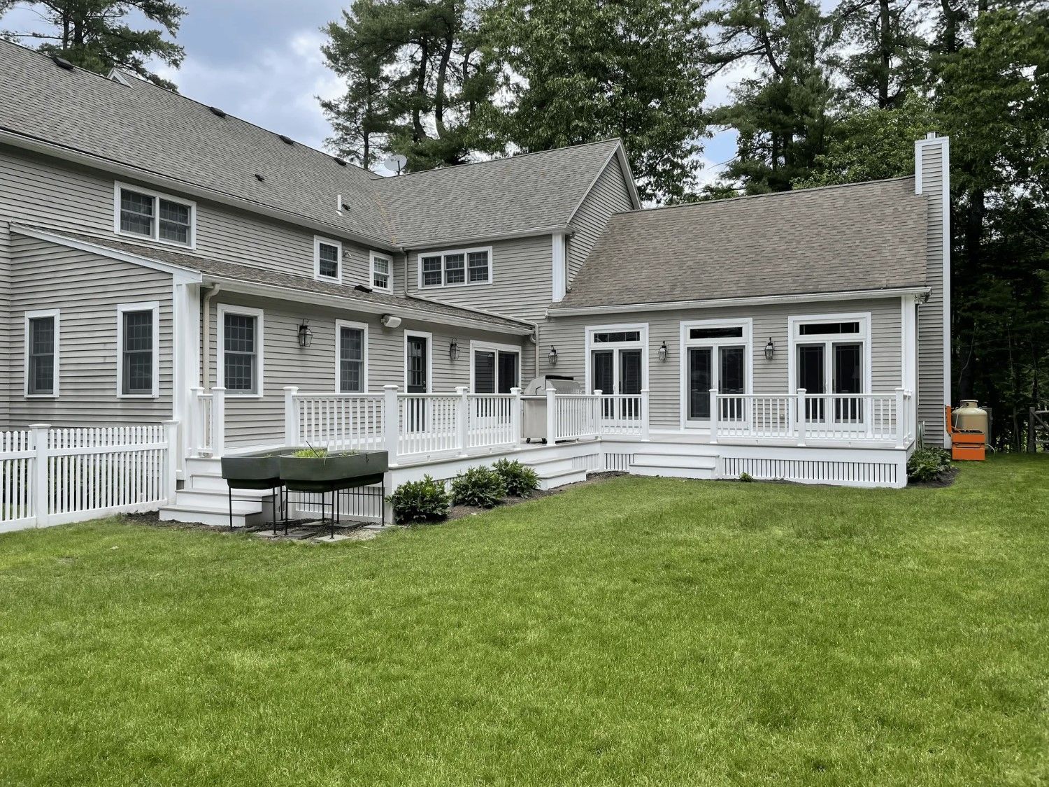 Back of a light gray house with a white deck, pergola, and green lawn. An orange propane tank stands on the right.