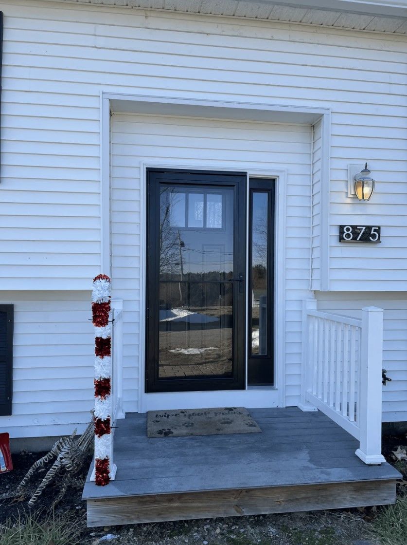 Black door with sidelight on a white house, gray porch, white railing, and the number 875.