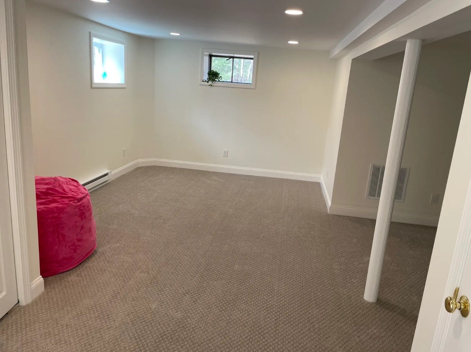Empty, carpeted basement room with two small windows, white walls, and a pink beanbag chair.