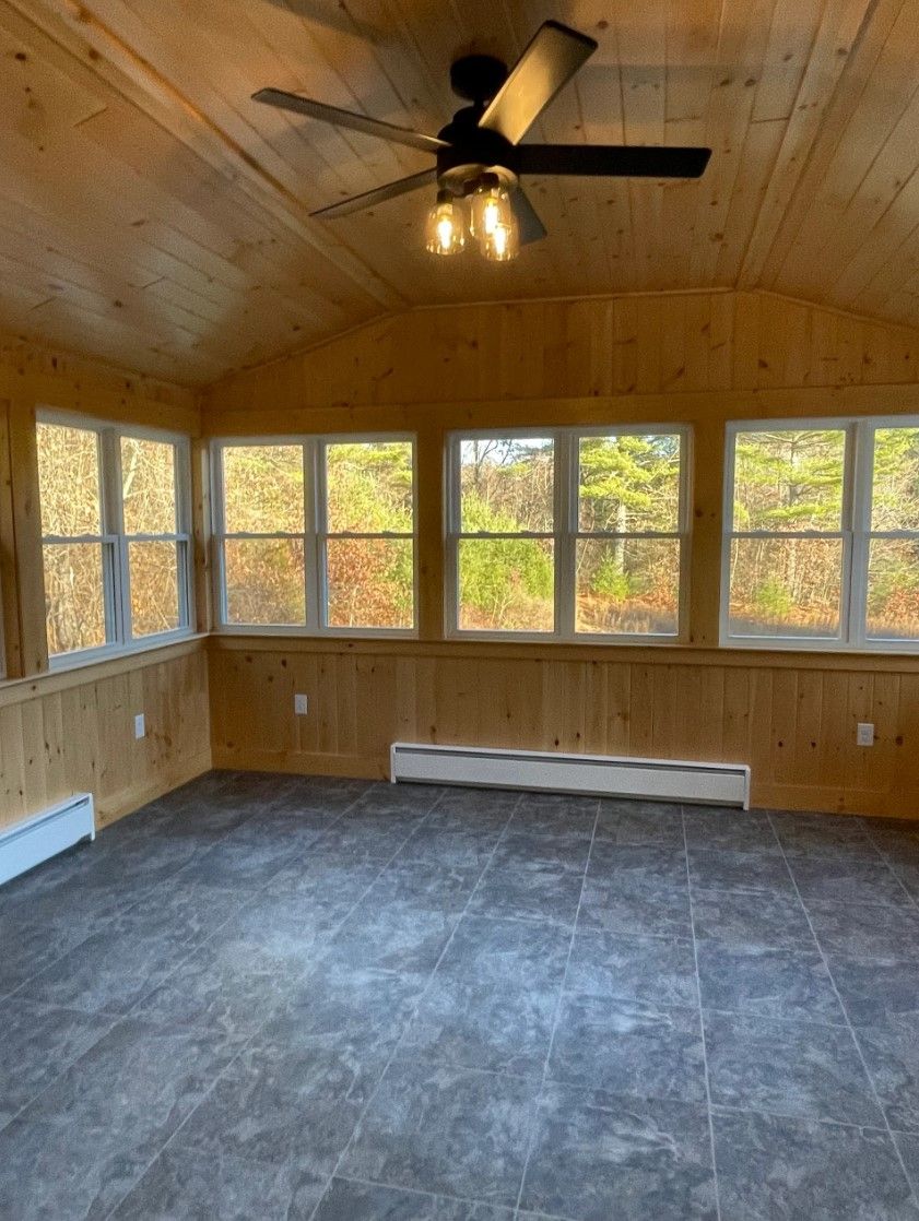 Sunroom with wooden walls, ceiling fan, large windows overlooking fall foliage, and gray tile floor.