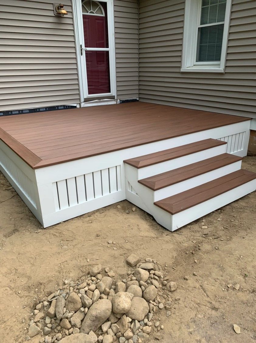 New brown composite deck with white trim and steps, beside a tan house with a red door.
