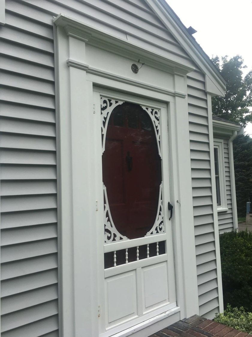 White screen door with ornate latticework and a red backing in a gray clapboard house.