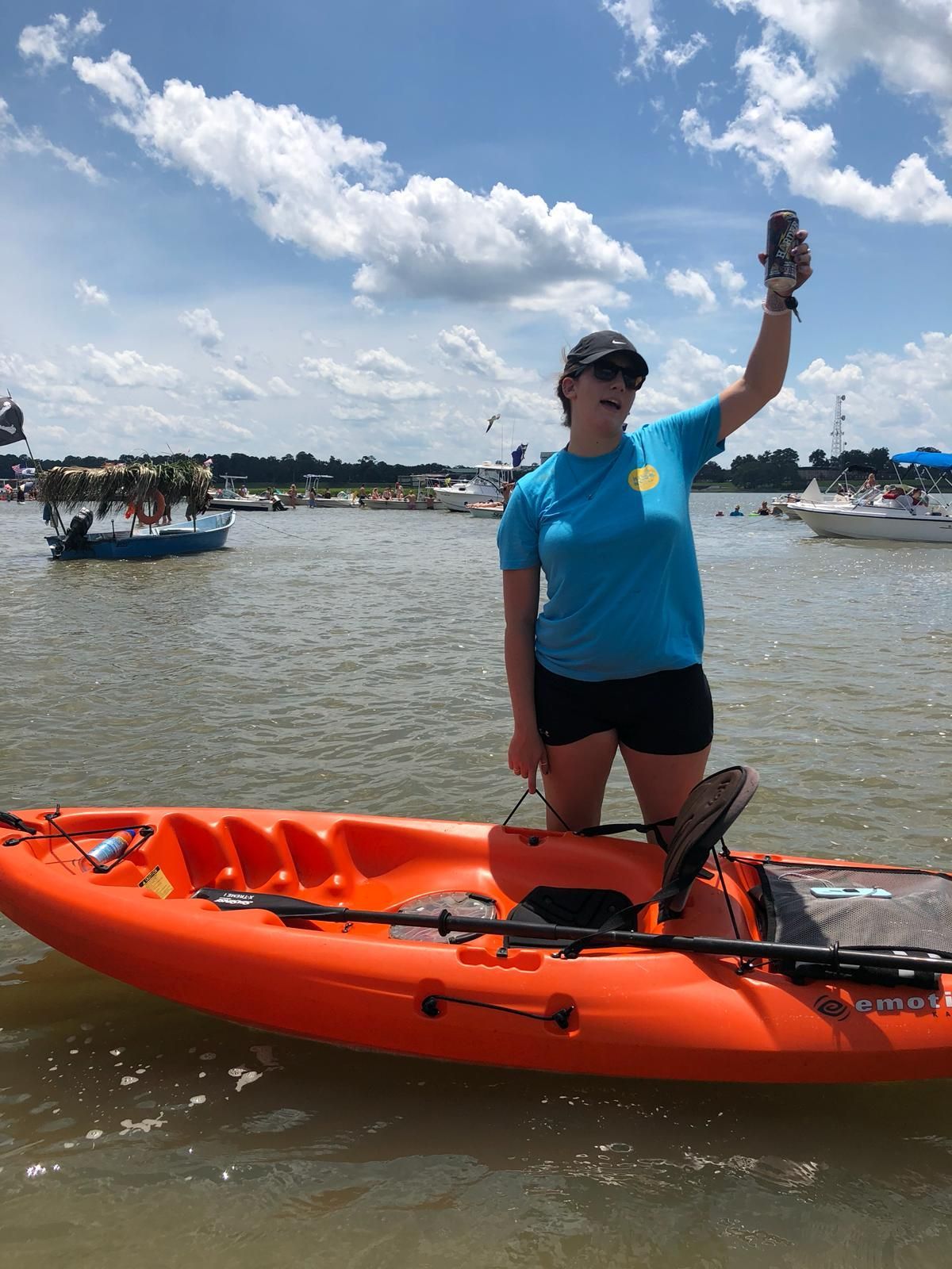 Sandbar party with woman on orange kayak raises a can in the air, smiling. Cloudy sky, other boats in the background.