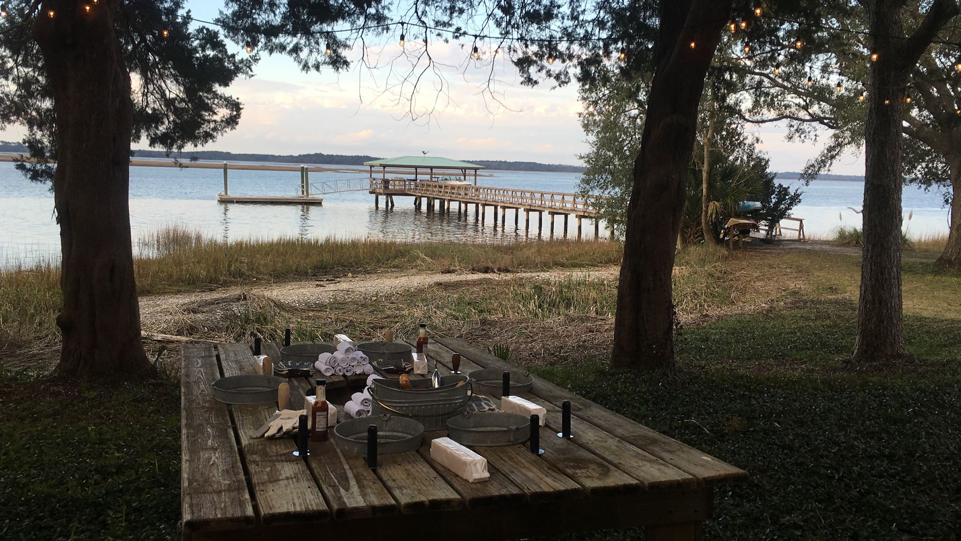 Picnic table set for a meal of oyster roast near a pier overlooking a body of water, trees in the foreground.