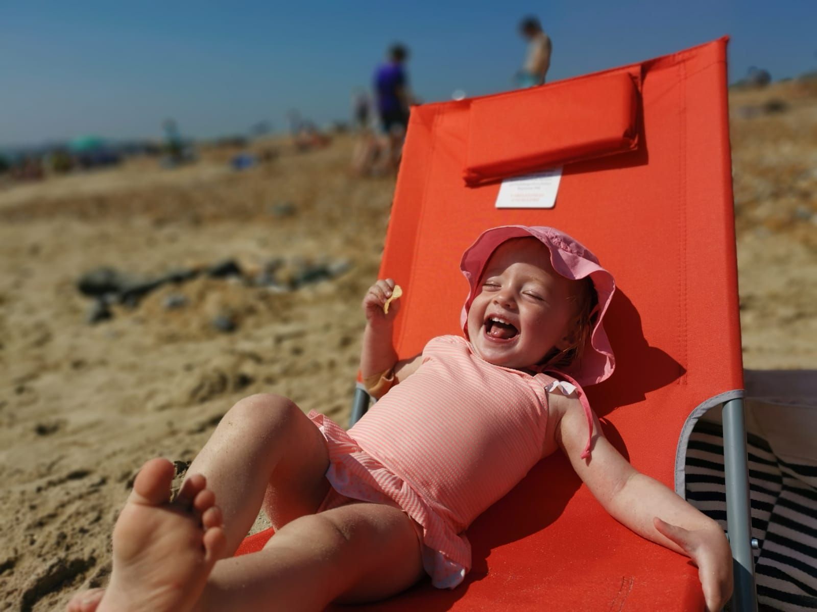 Child laughing while reclining in a bright orange beach chair, wearing a pink hat and swimsuit. Beach setting.