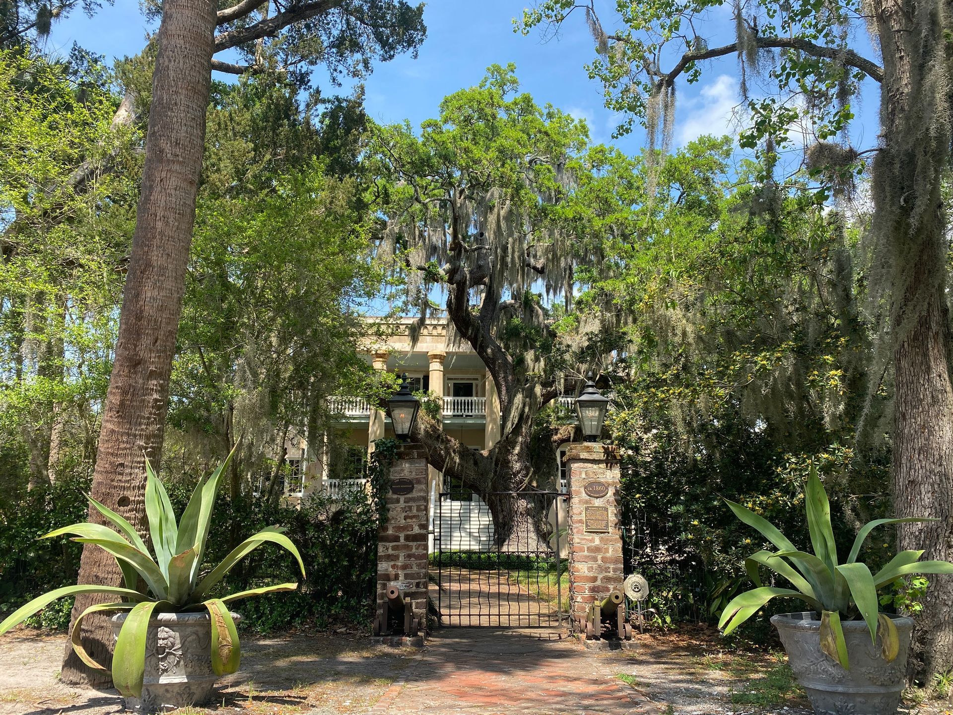 Stone gate leads to a historic white house with a porch, framed by trees and plants with Spanish moss.