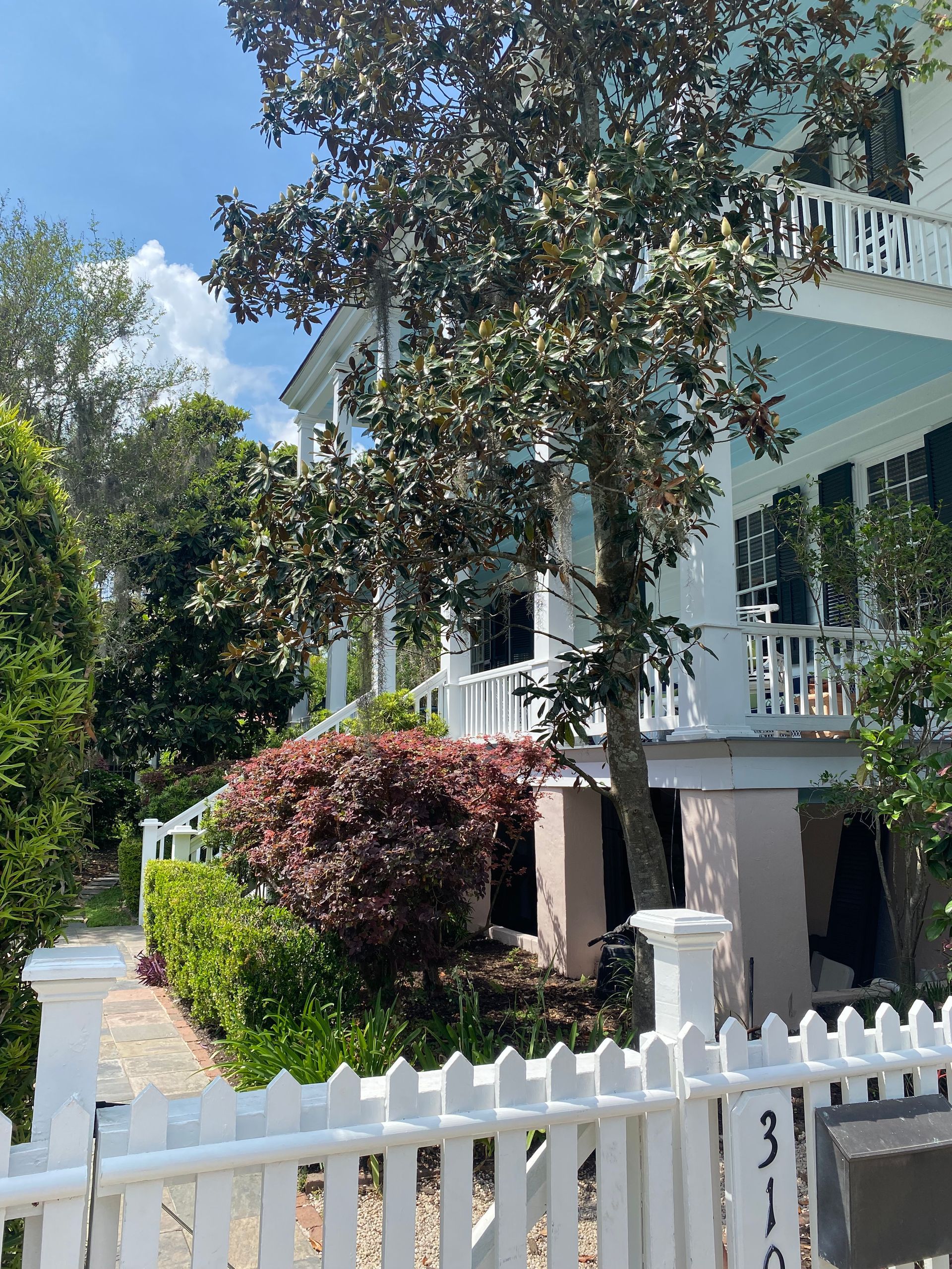 White house with balcony, light blue ceiling, and white picket fence. Green and red bushes in front.