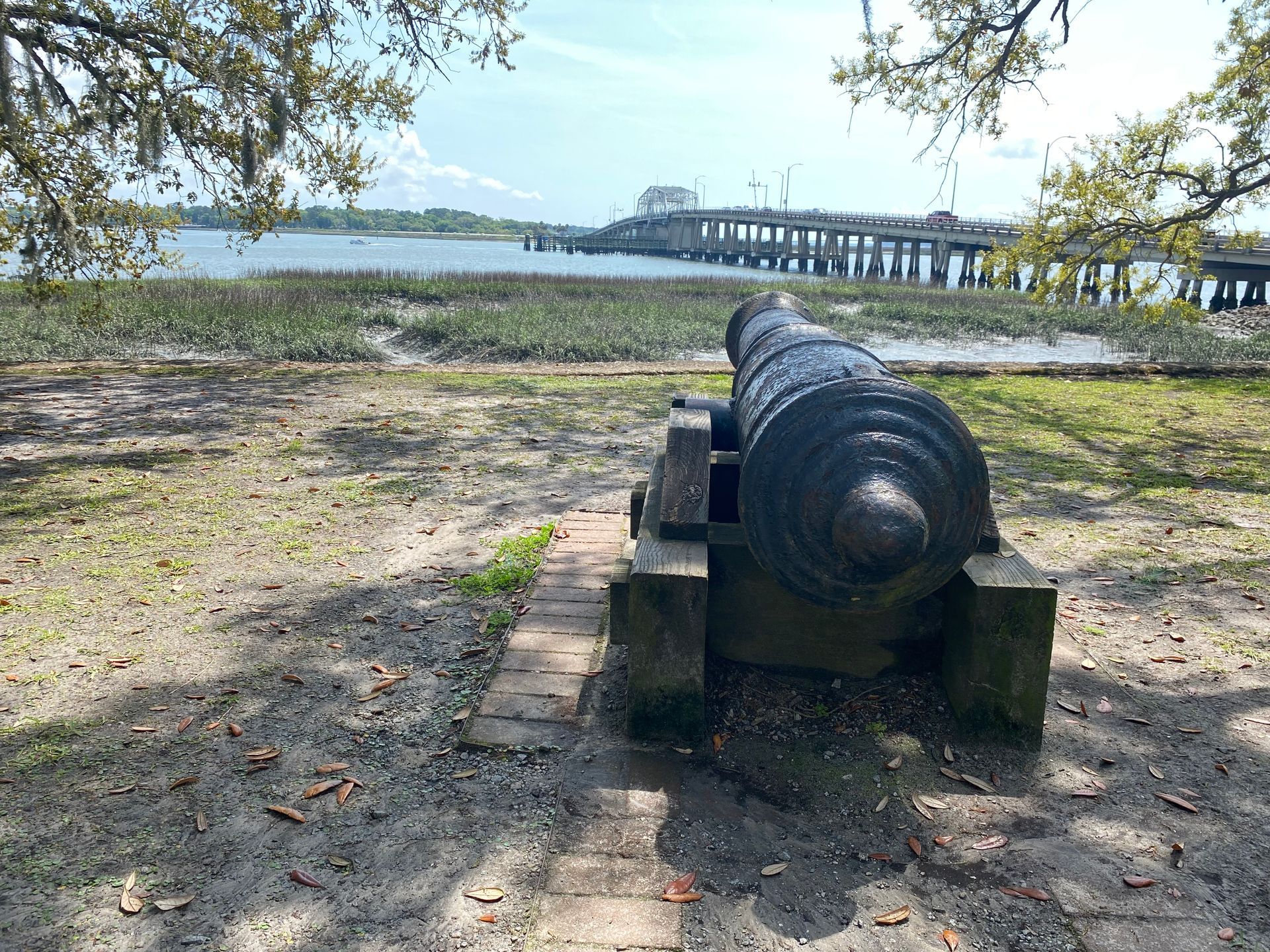 Old cannon facing water, with a pier in the background, under a blue sky.