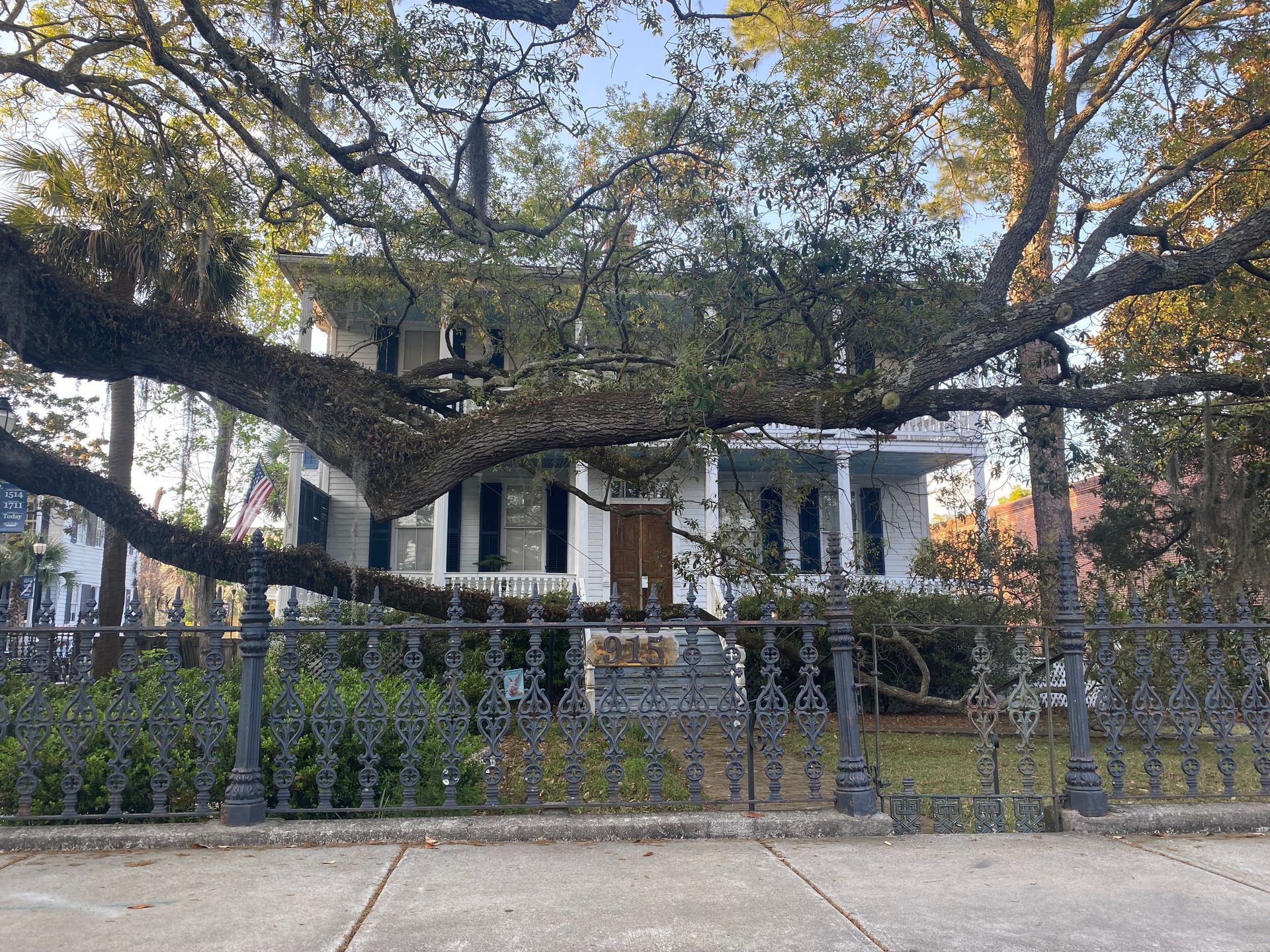 Lowcountry white house with a porch behind a wrought iron fence, framed by large tree branches.