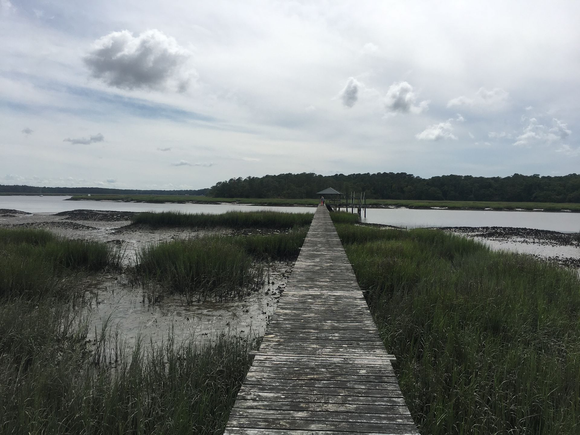 Wooden pier extending over marshy water towards a gazebo, under a cloudy sky.