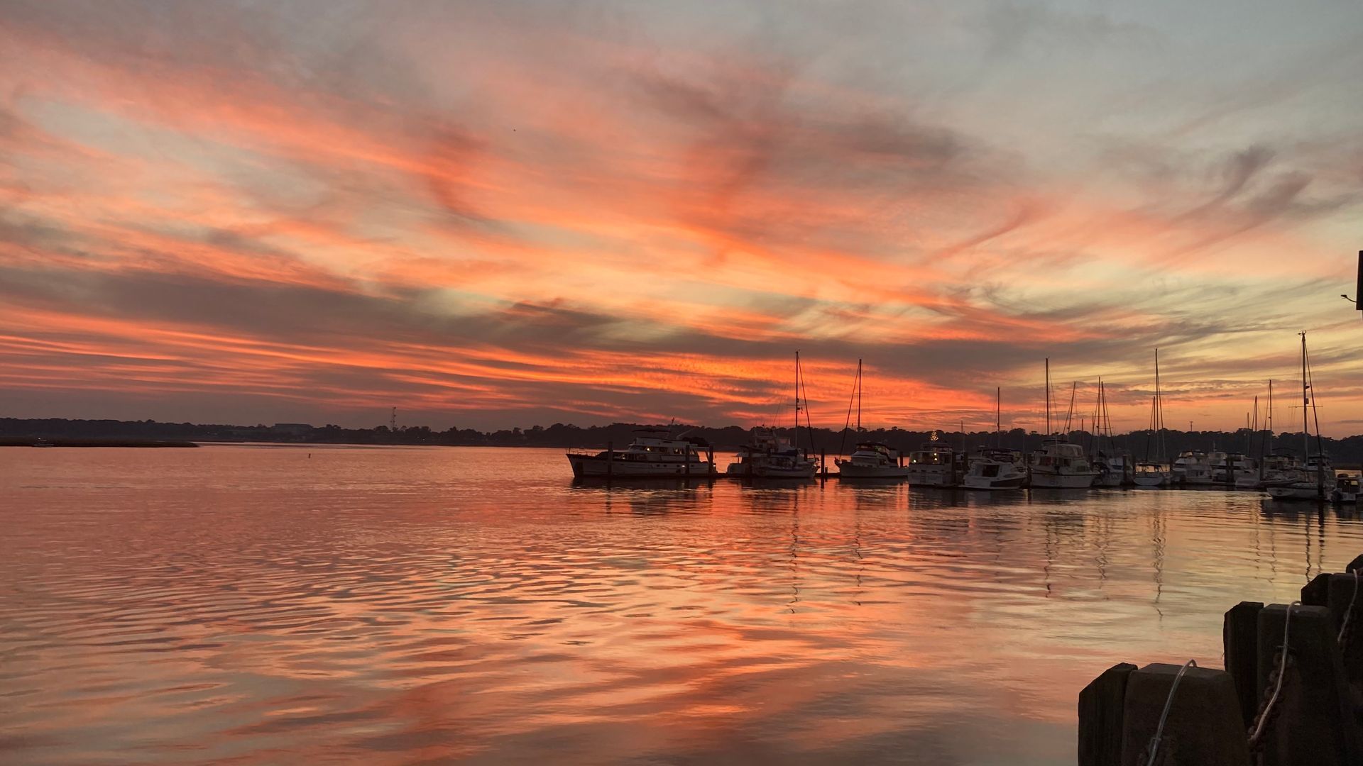 Vibrant orange and pink sunset reflecting on calm water, sailboats docked in a harbor.