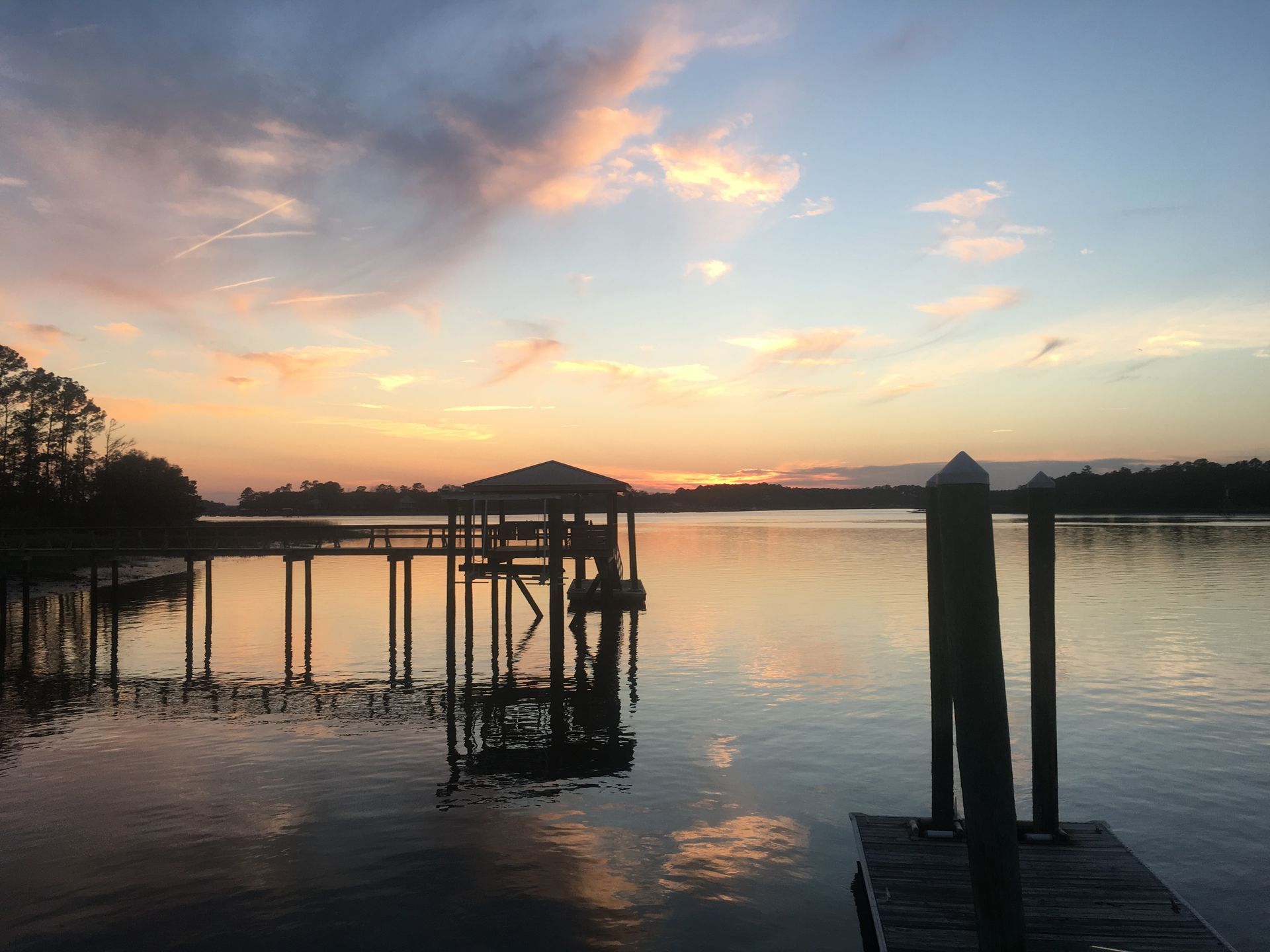 Sunset over calm water with a dock, fishing pier and posts. Reflecting colors: orange, pink, and blue.