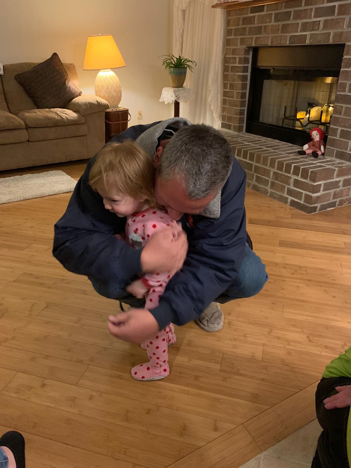 Man hugging a toddler wearing pink pajamas indoors, near a fireplace and couch.