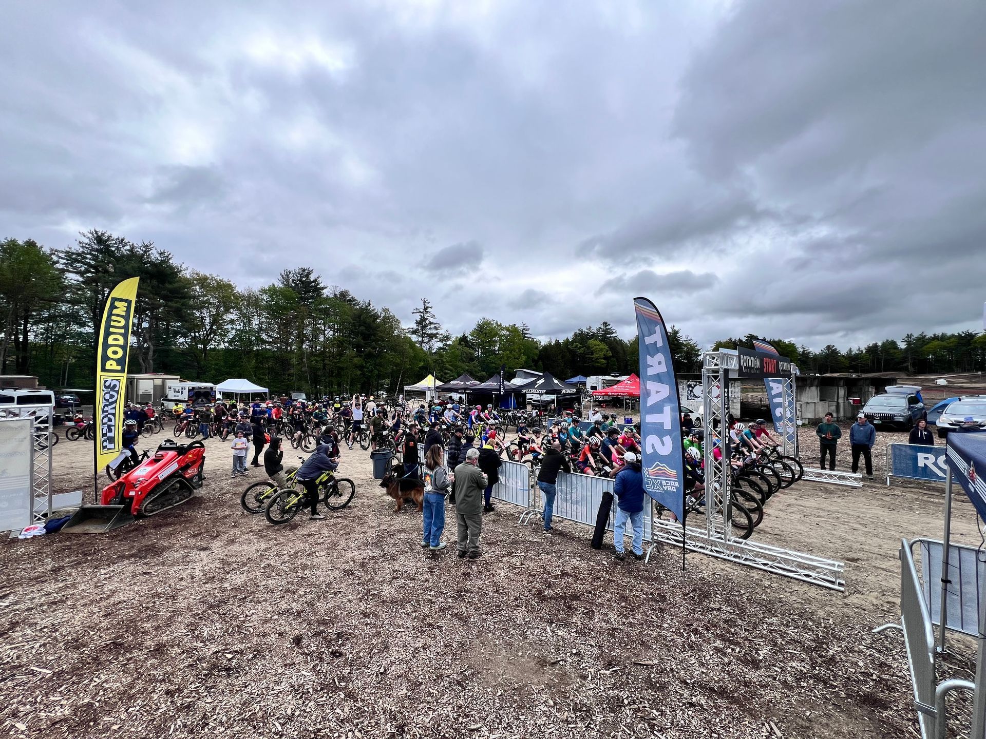 A crowd gathers at an outdoor event with flags and vehicles under an overcast sky.