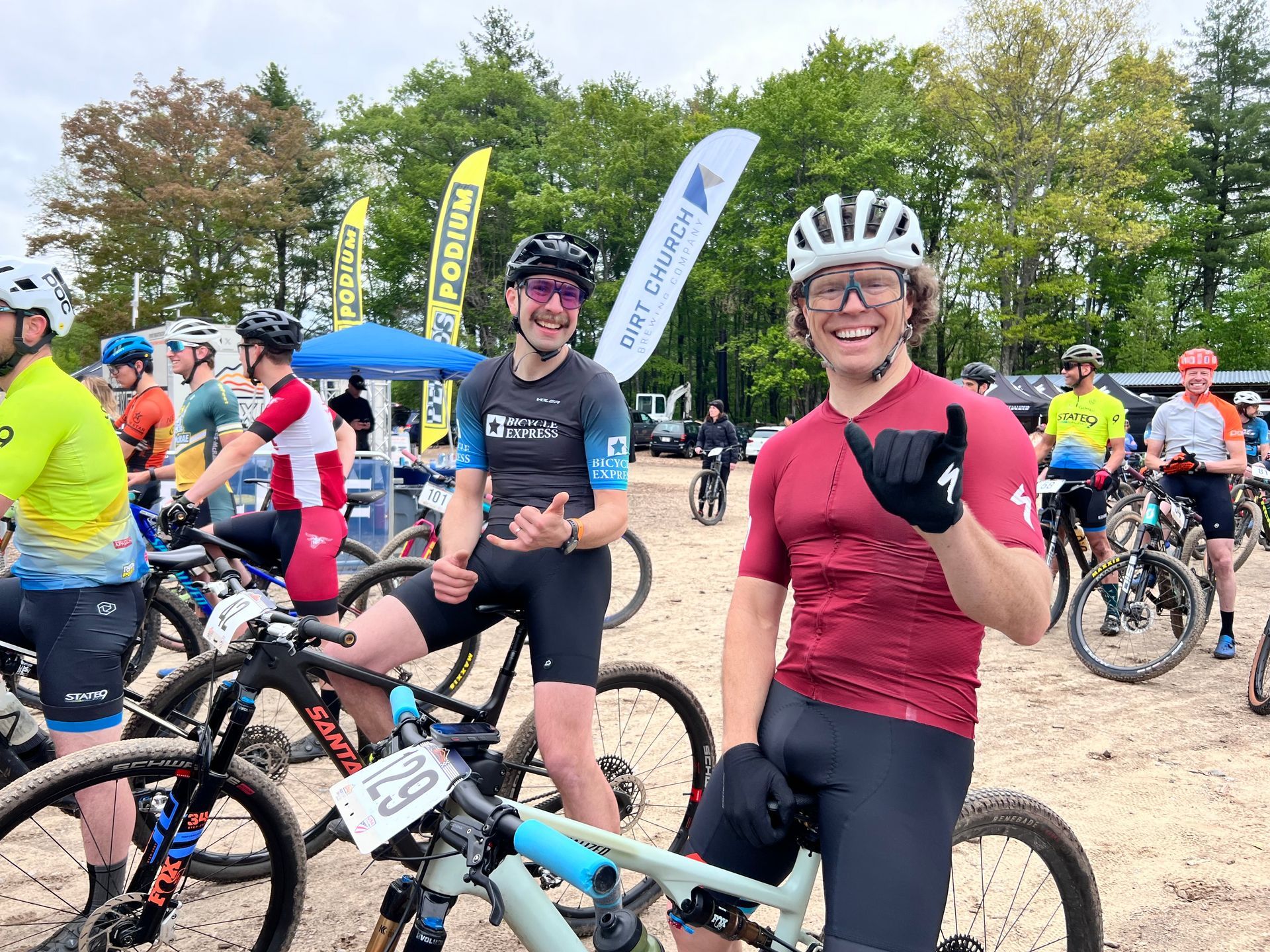 Cyclists on bikes at outdoor event; two smiling, giving hand gestures.