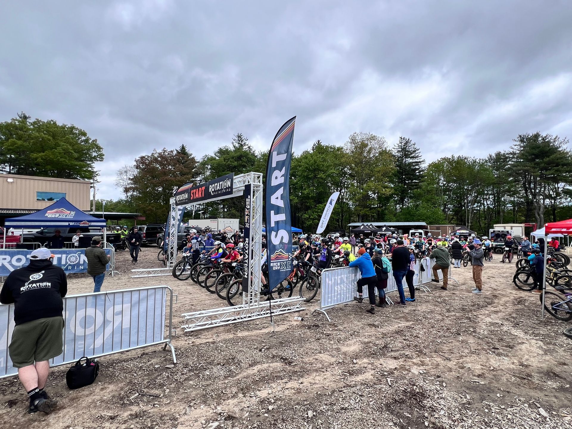 Bicycle race starting line; riders on bikes behind barriers. People watch near tents under a cloudy sky.