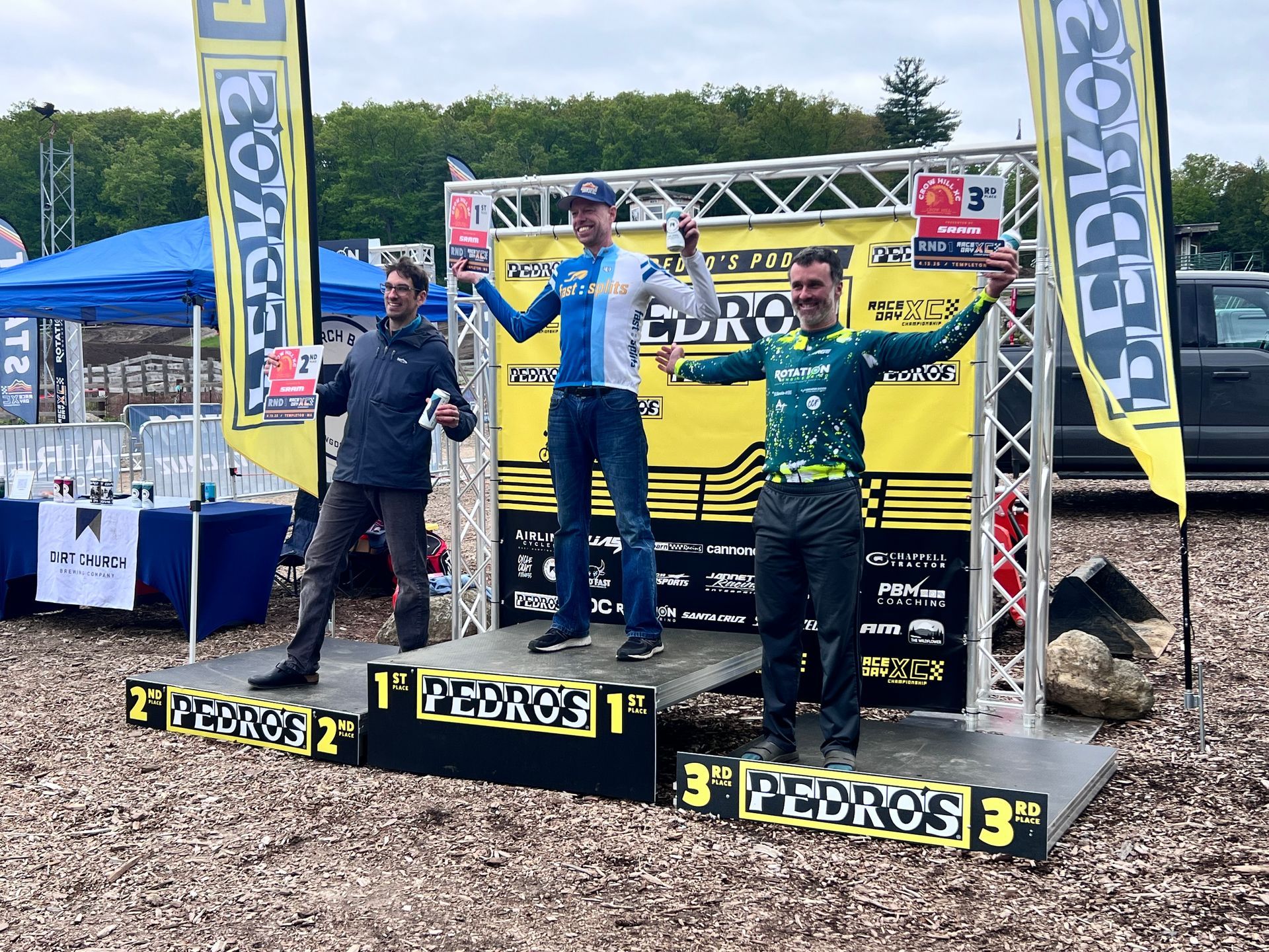 Three people on podium, celebrating a cycling event. 1st place in center, arms raised, 2nd and 3rd on sides.
