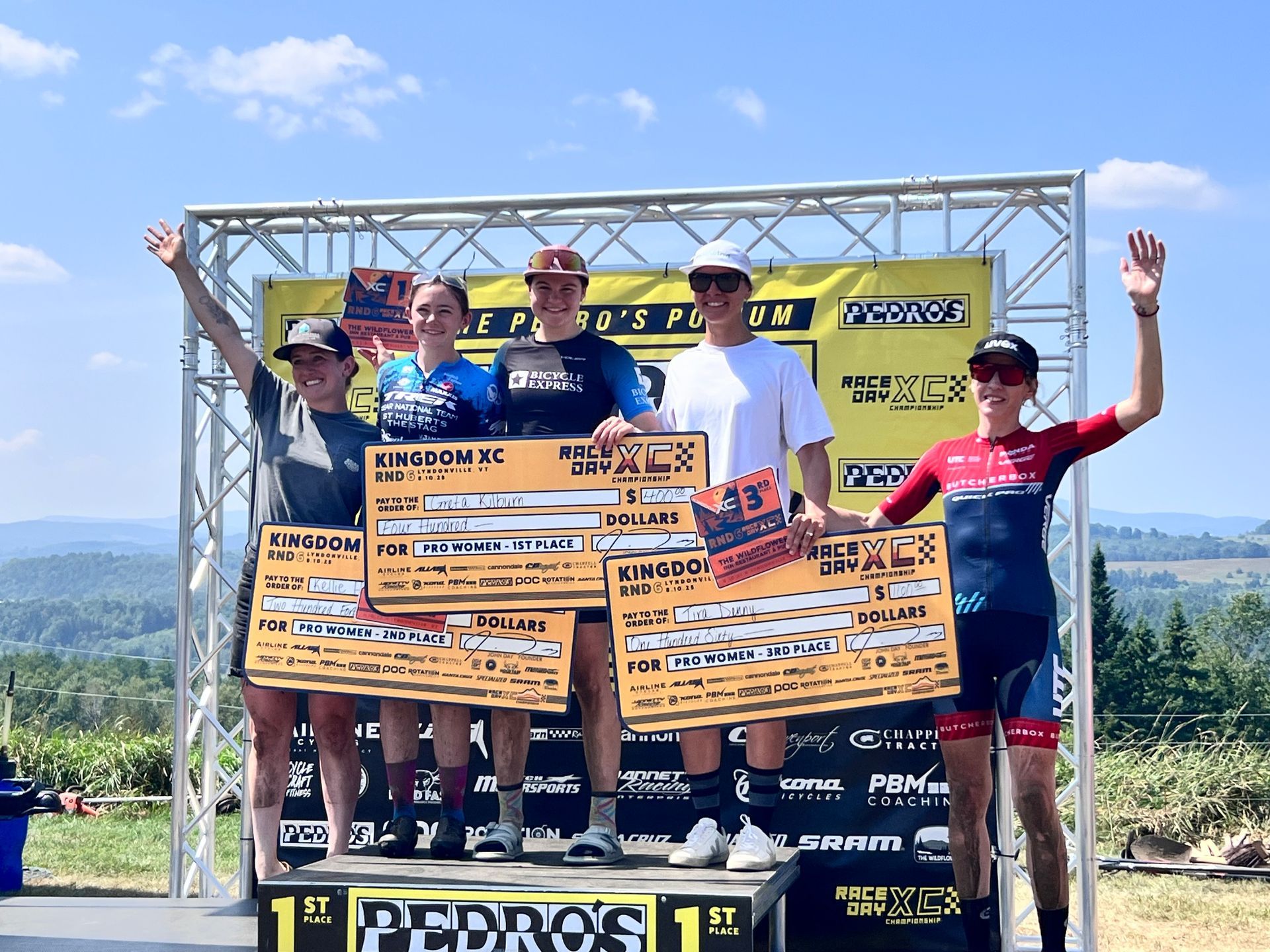 Cyclists on podium, holding oversized checks. Sunny outdoor setting with a banner and cheering.