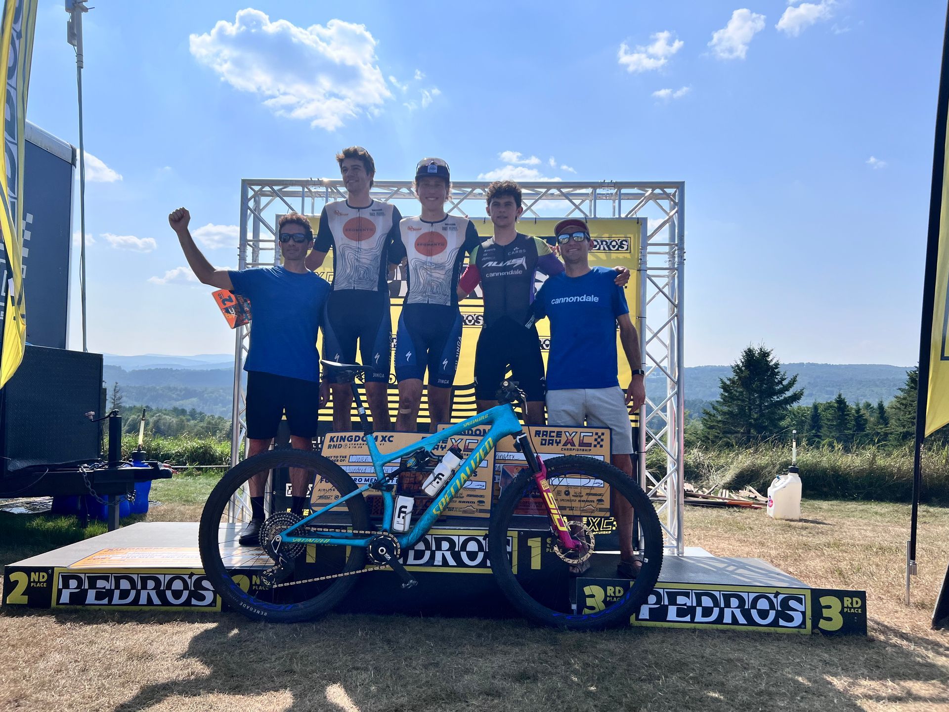 Mountain bike on podium with winners, outdoors. Riders on podium, blue sky, sponsors' banners.