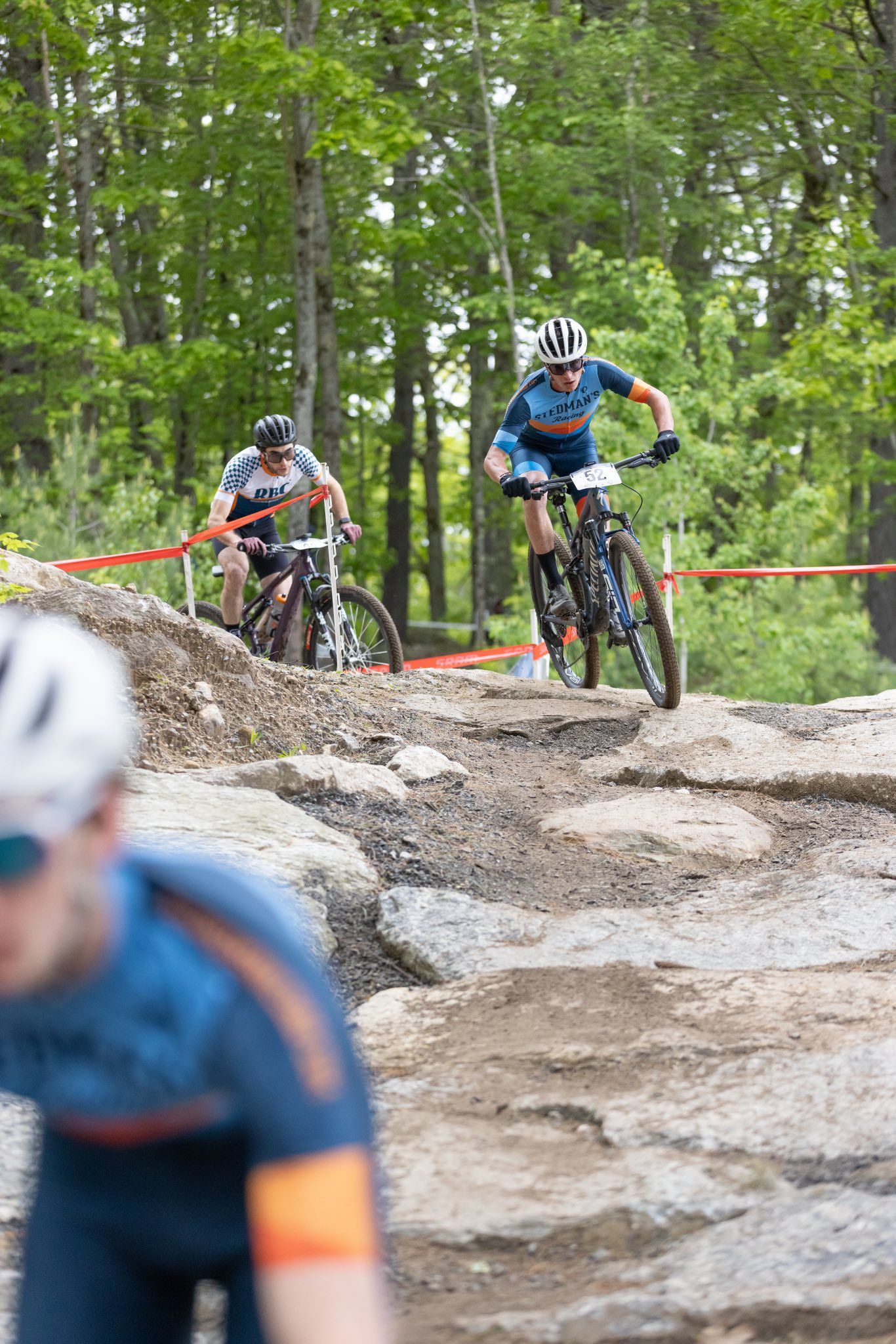 Mountain bikers racing on a rocky trail in a wooded area. One rider in blue leads.