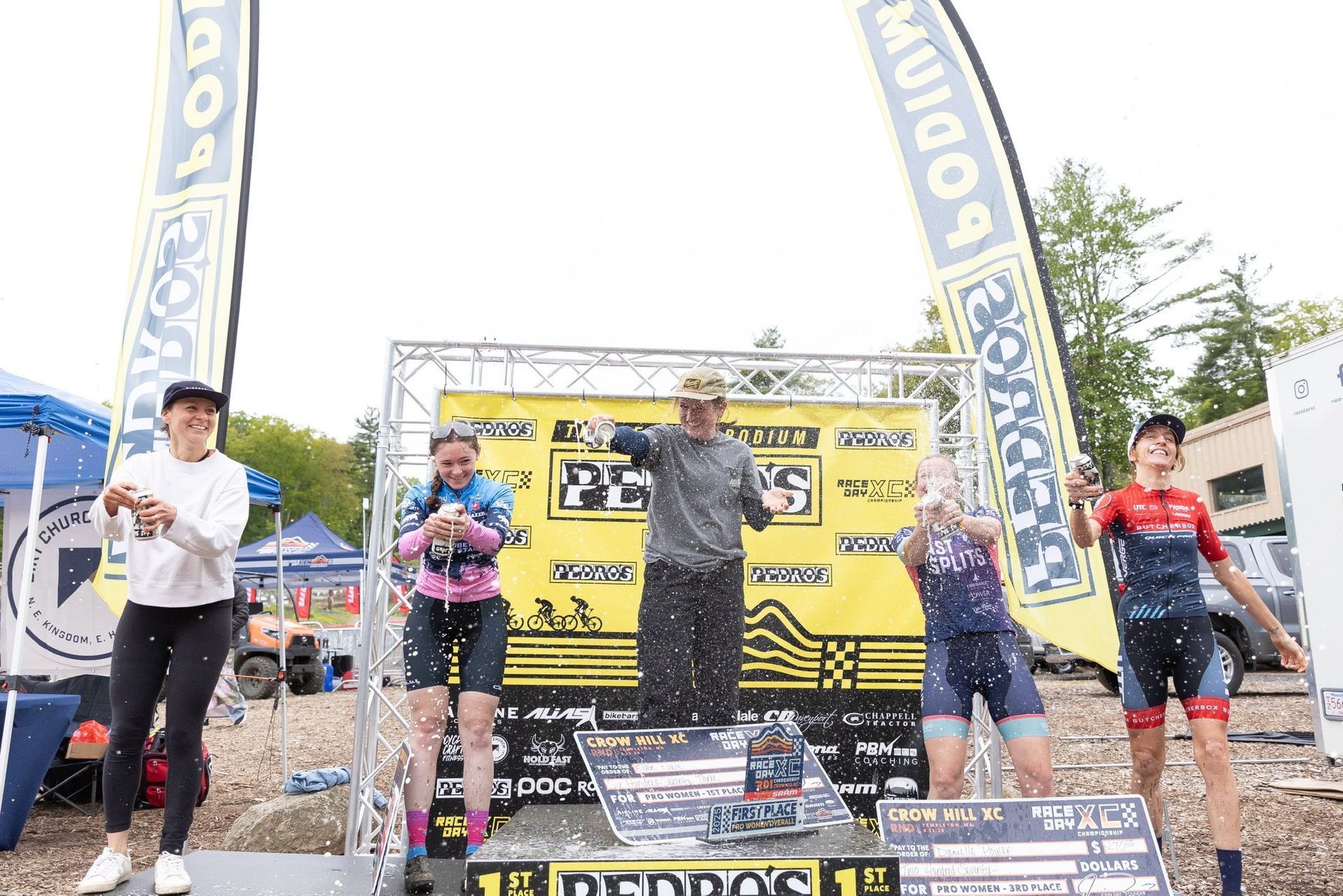 Cyclists on podium, sprayed with confetti. Event banner behind them.