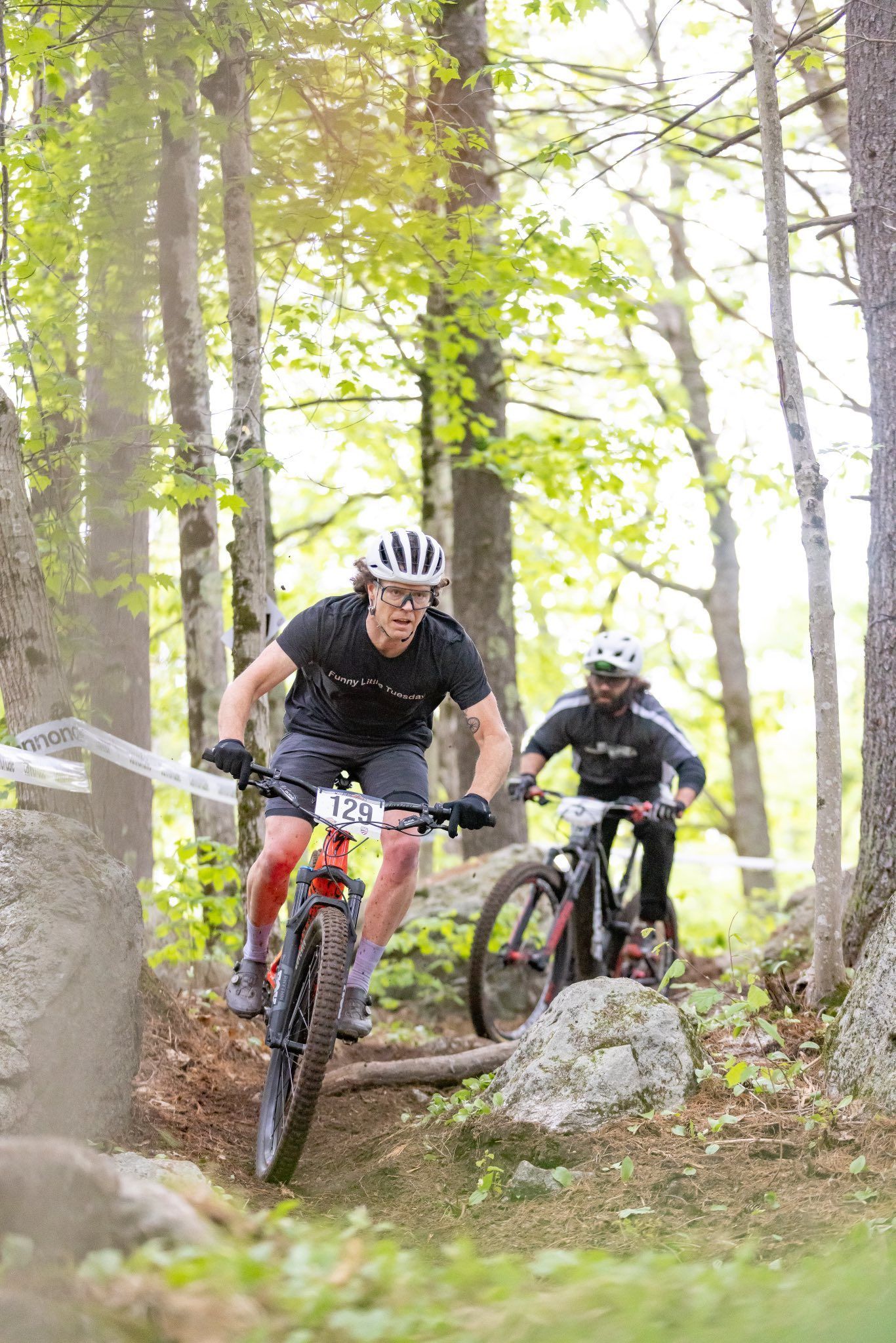 Two mountain bikers racing on a wooded trail. One leads, both wear helmets and ride through trees.