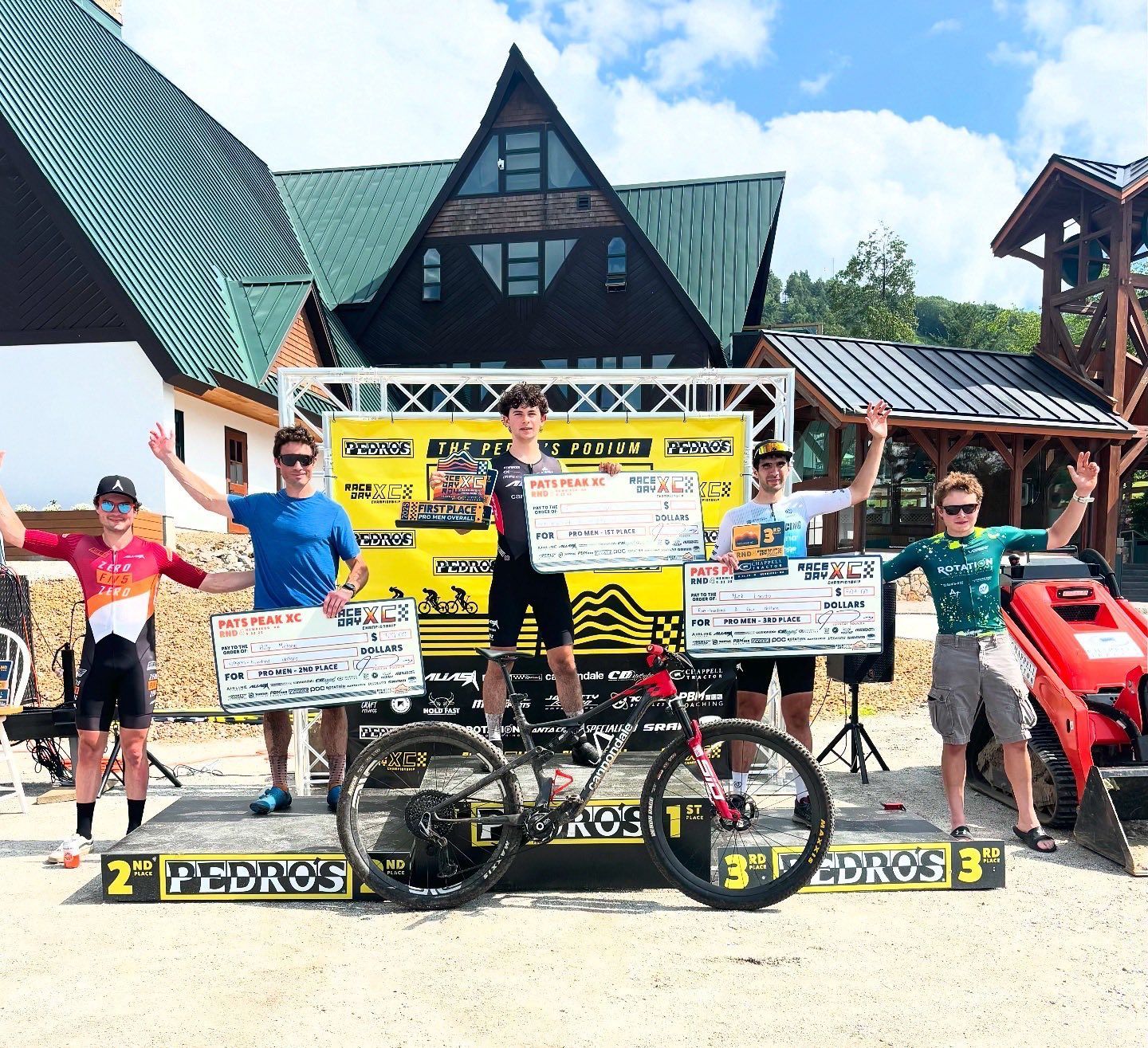 Cyclists on podium, holding prize checks, after a mountain bike race. Yellow banner, sunny day.