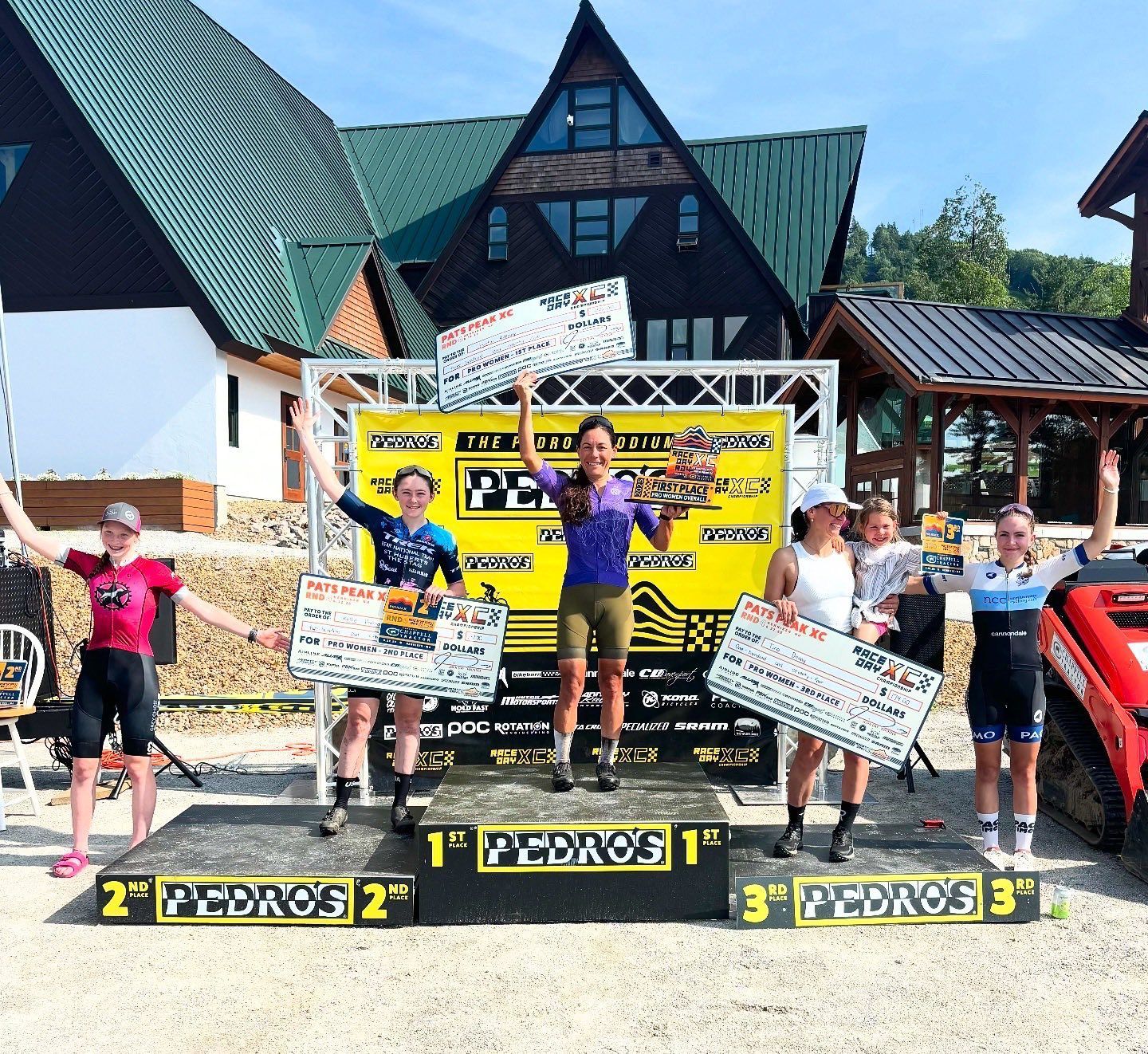 Mountain bike racers on a podium, holding awards. Green and yellow event banner, outdoor setting.