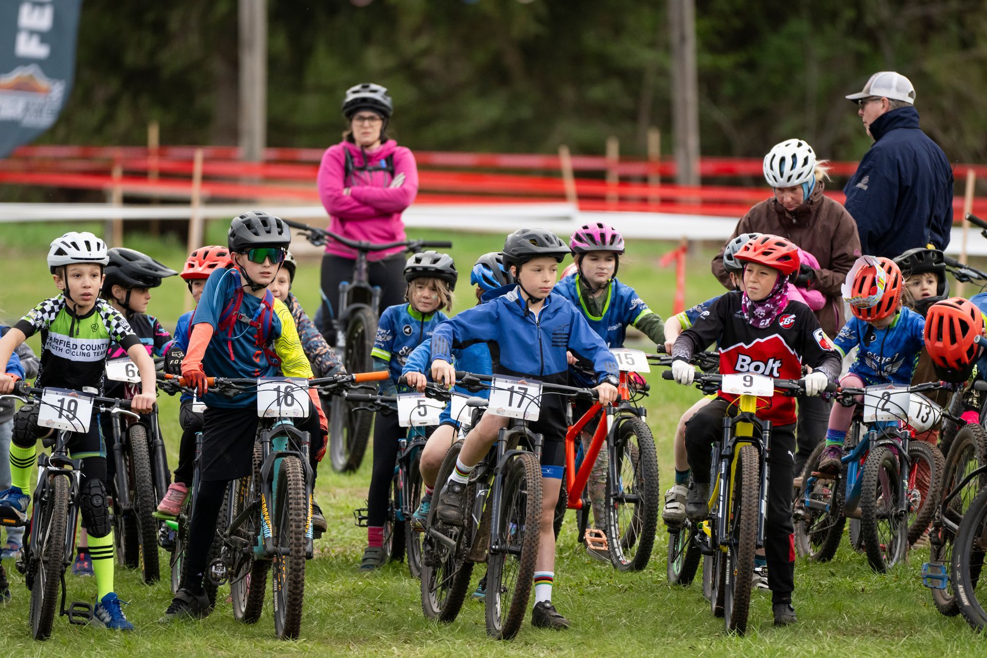 Children on bikes lined up at the start of a mountain bike race outdoors. People in background.