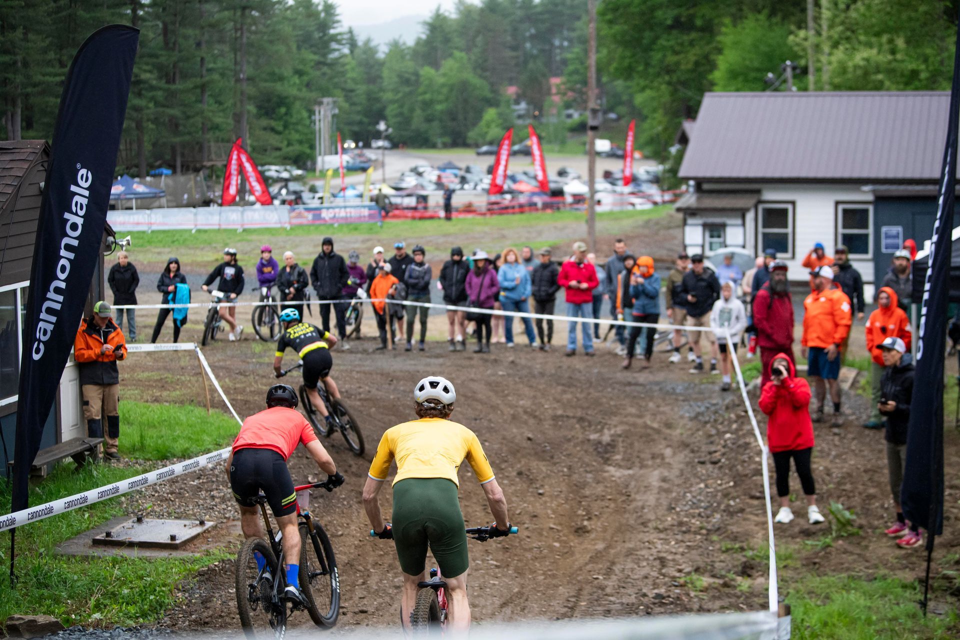Mountain bike race: Cyclists on dirt track with spectators, banners, trees, and buildings in the background.