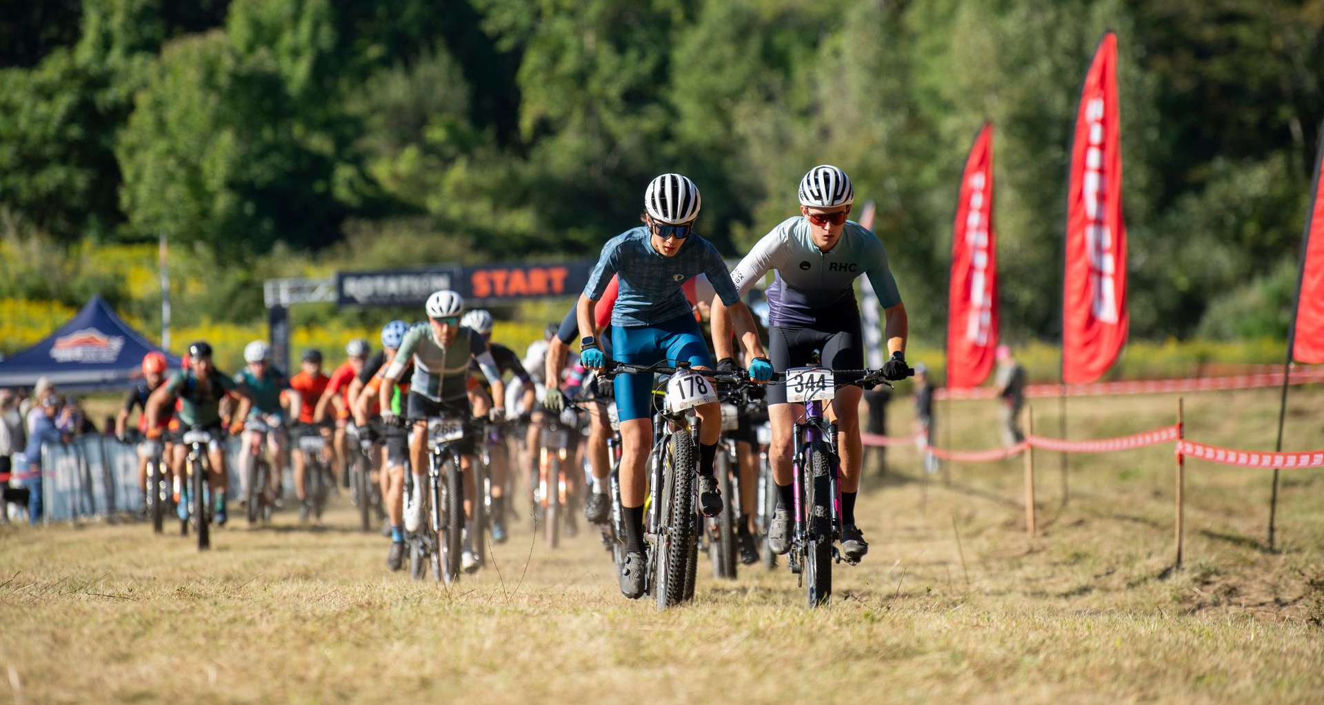 Mountain bikers racing on a dirt path; start line in background, sunny day.