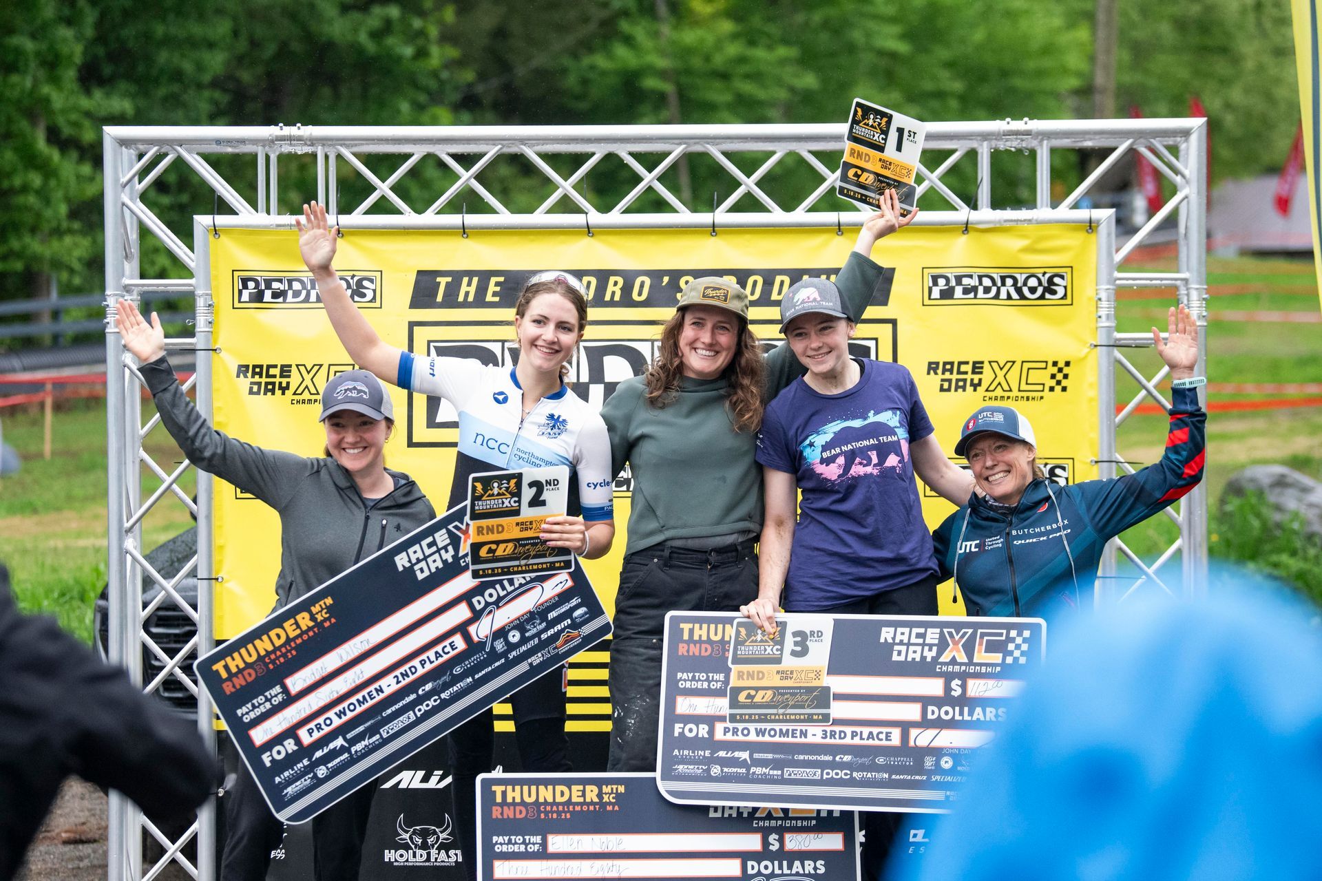 Winners on a podium holding awards at an outdoor event, with a yellow banner and metal scaffolding in the background.