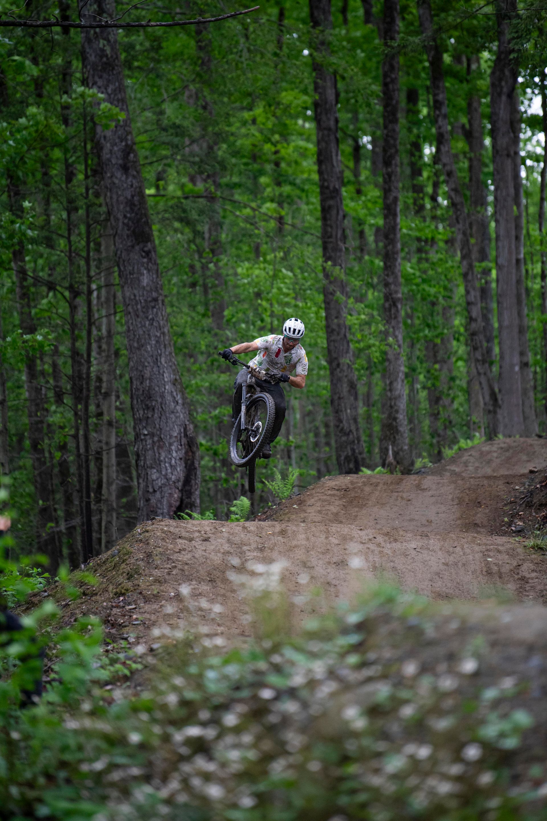 Mountain biker in mid-air over a dirt jump, surrounded by a forest of green trees.