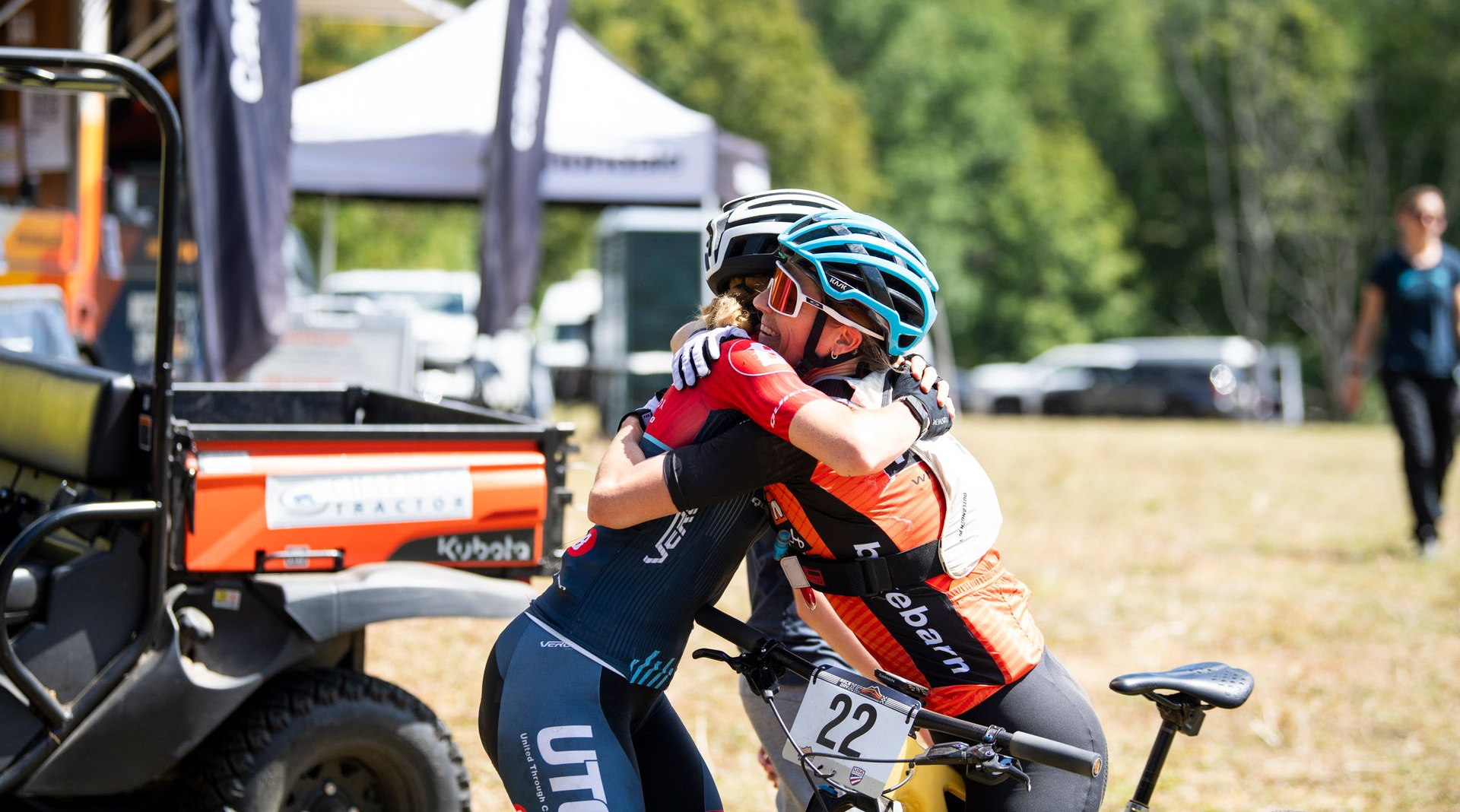 Two cyclists in helmets embrace, post-race. One wears blue, the other orange. Outdoors, sunny day.