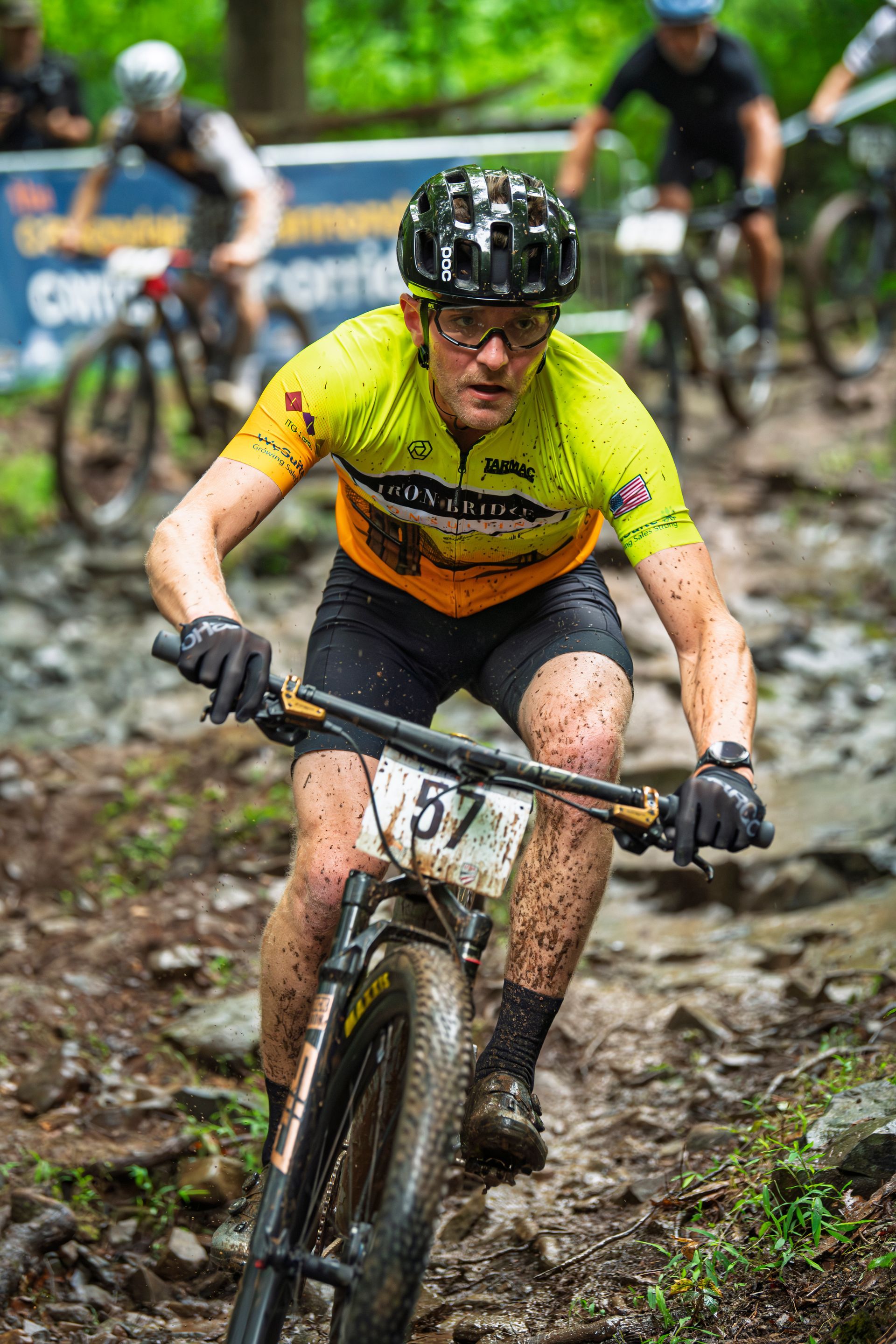 Mountain biker (#57) on a muddy trail, wearing a yellow jersey, helmet, and glasses.