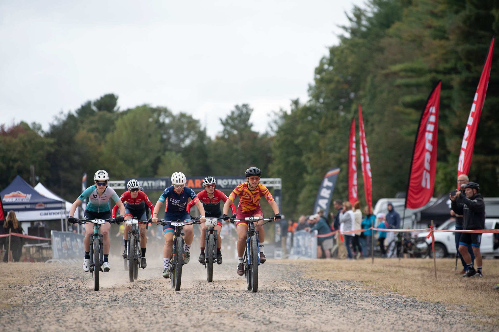 Mountain bikers racing on a dirt road, trees and tents in the background, red flags along the side.