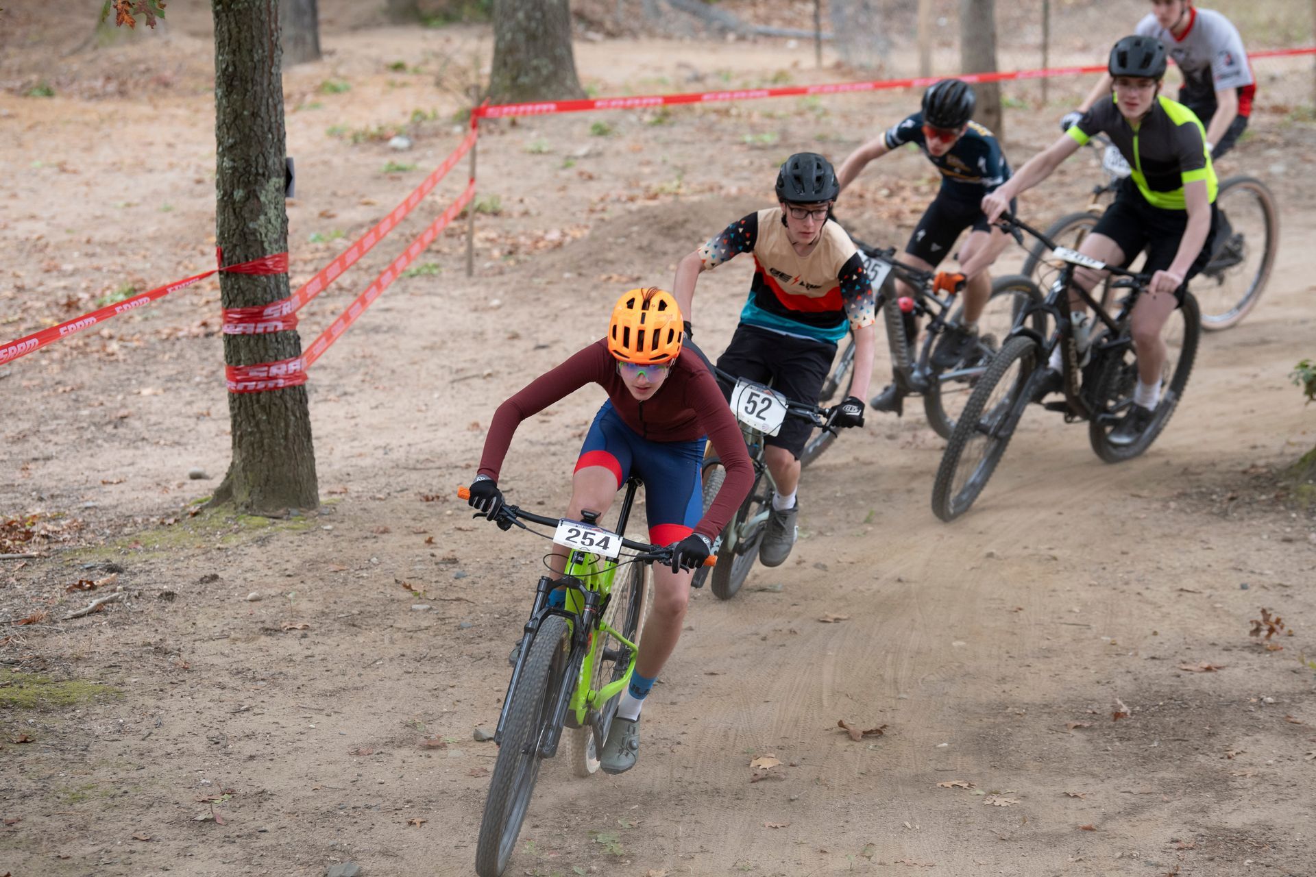 Mountain bikers racing on a dirt path in a wooded area. The rider in front is wearing an orange helmet and blue and red shorts.