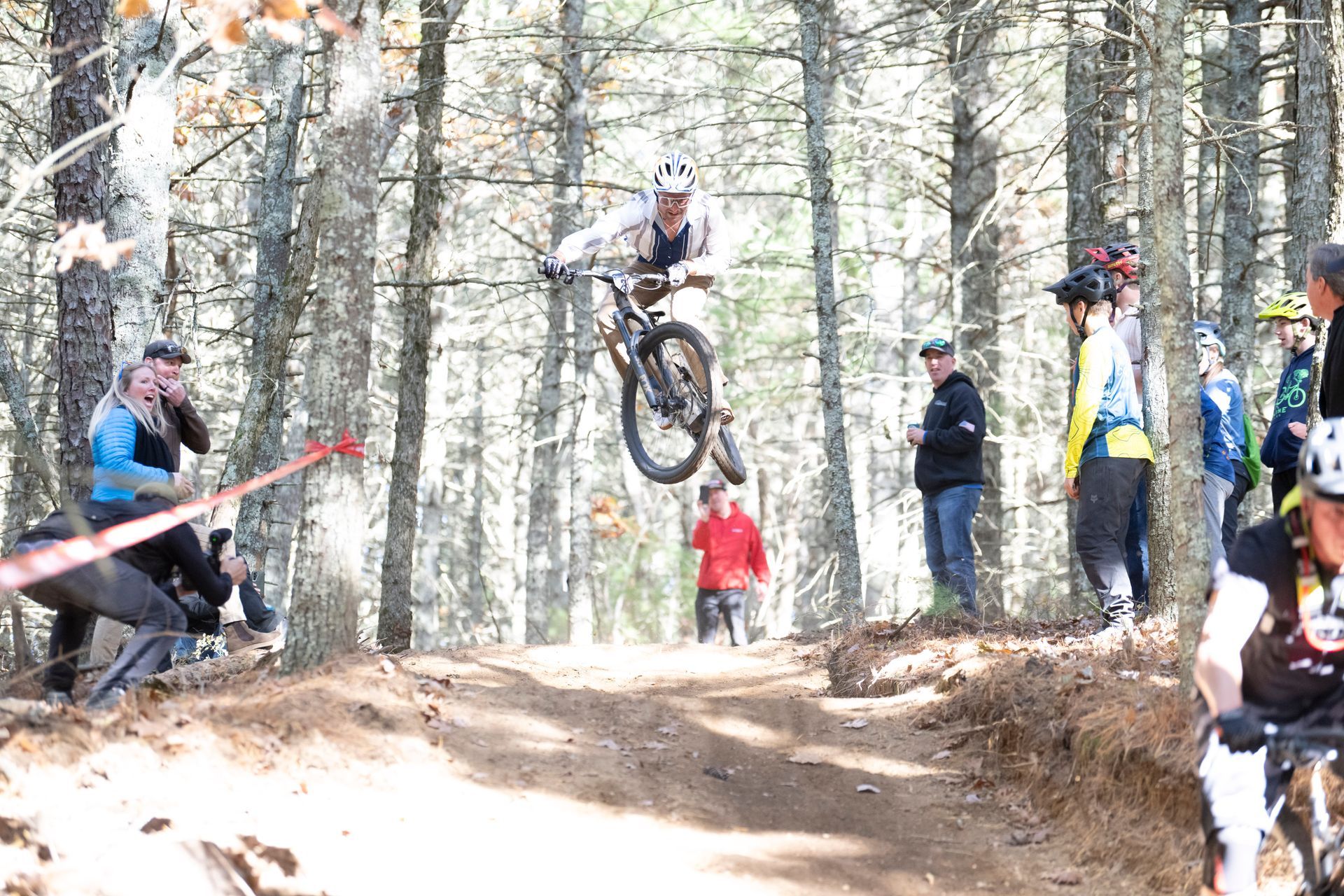 Mountain biker in mid-air over a dirt jump, surrounded by trees and spectators.