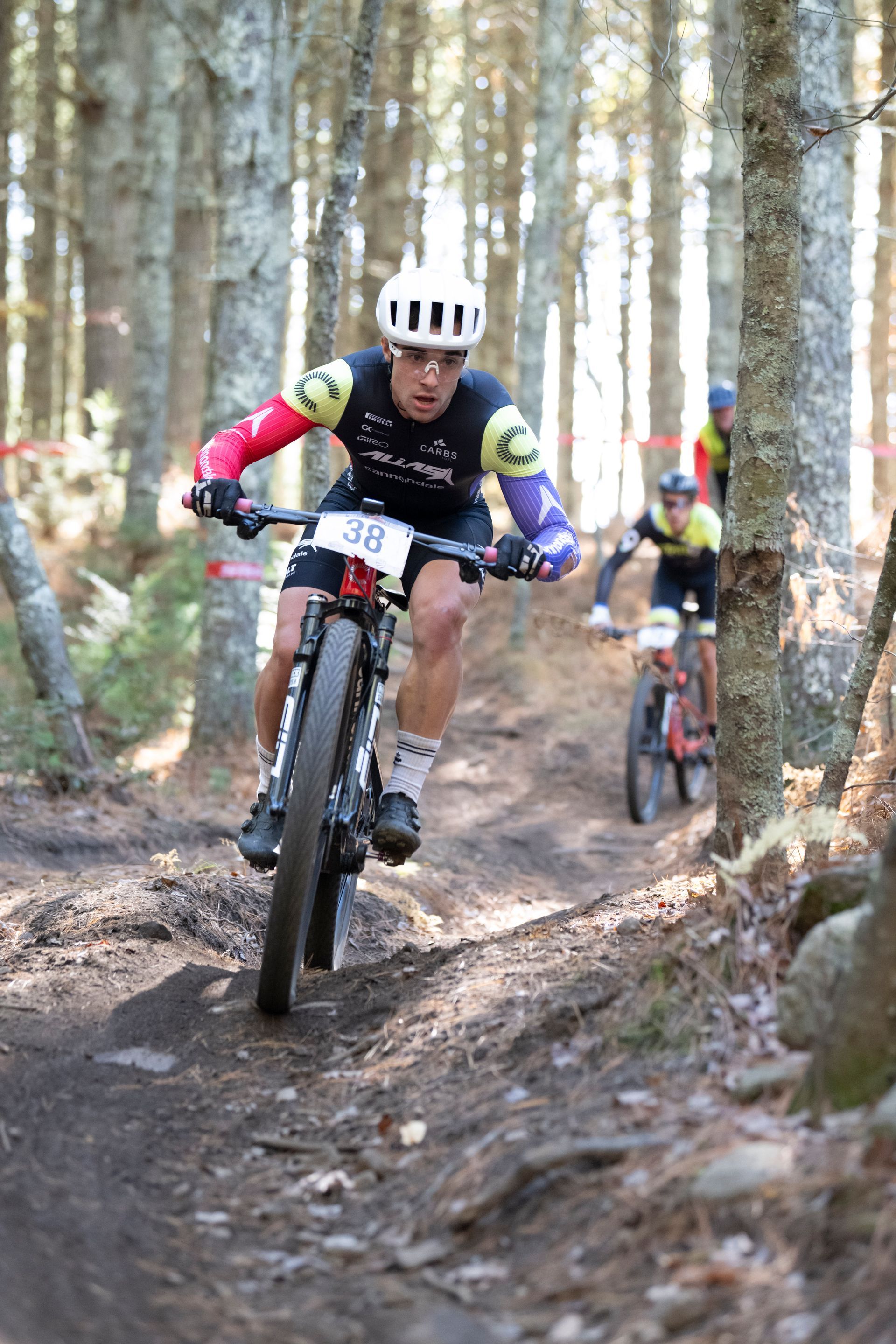 Mountain biker racing on a wooded trail. The rider wears a helmet and colorful jersey. Another biker follows.