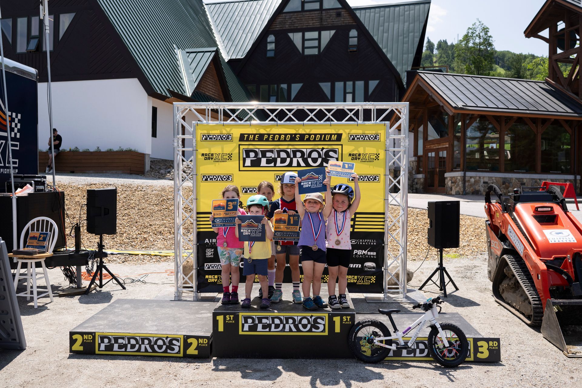 Children stand on a podium, holding trophies. A yellow banner reads “PEDROS.” A small bike sits at the 3rd place.