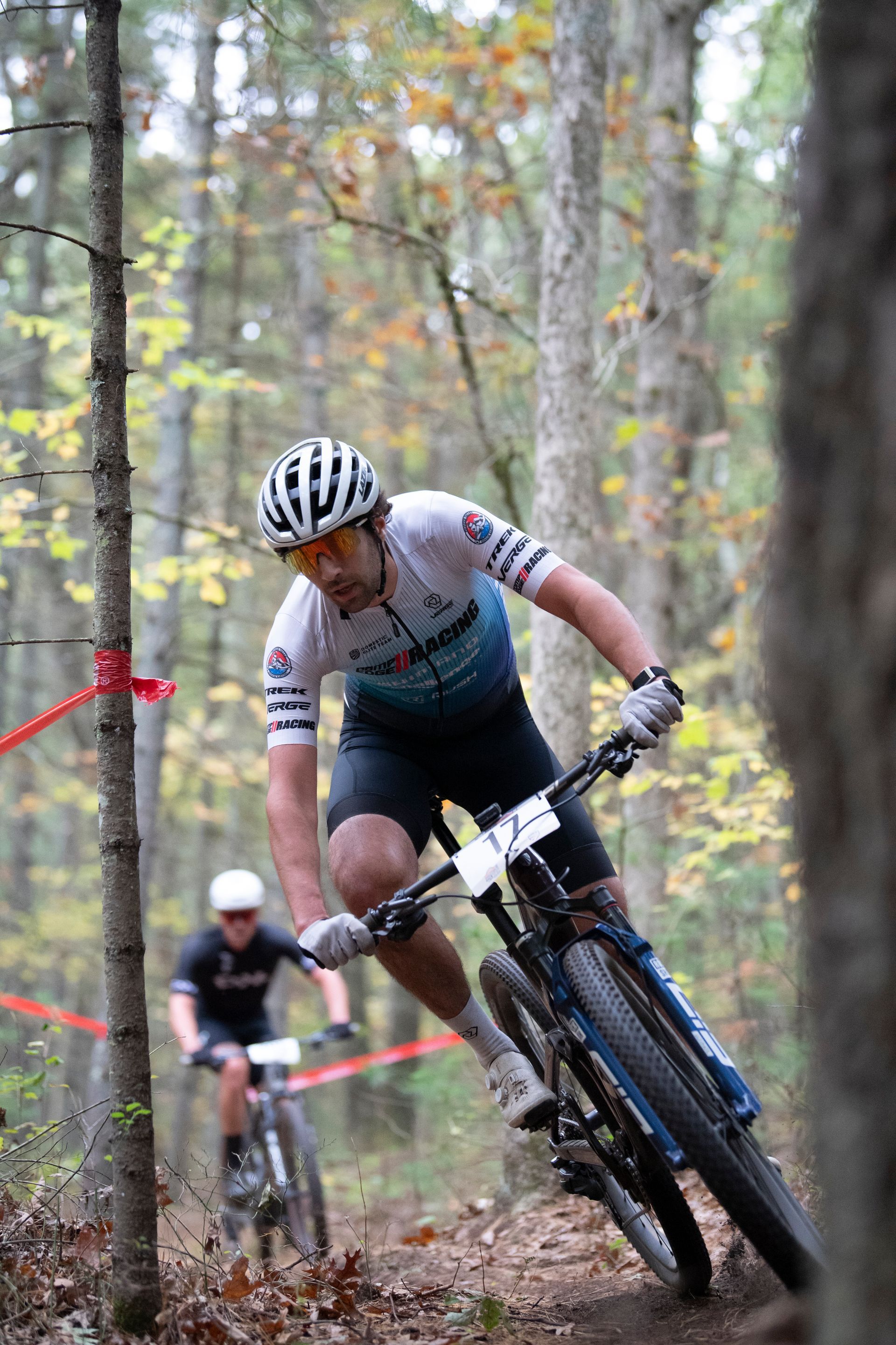 Mountain biker in a forest turns a corner, leading another cyclist in a race. Red tape and trees are in the background.